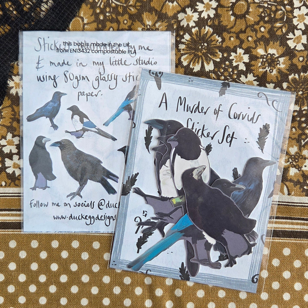 Two sticker sets lie next to each other one showing the front and the other showing the back of the set. Behind the sticker sets is a brown patterned fabric.