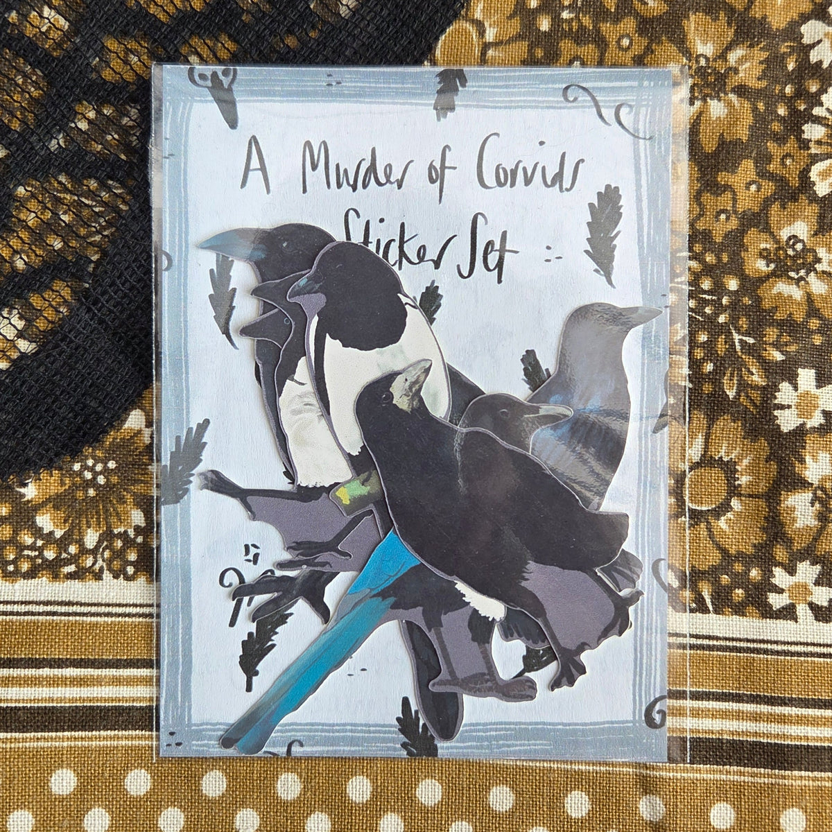 A sticker set sits on a brown patterned fabric showing the corvid stickers inside of it. 