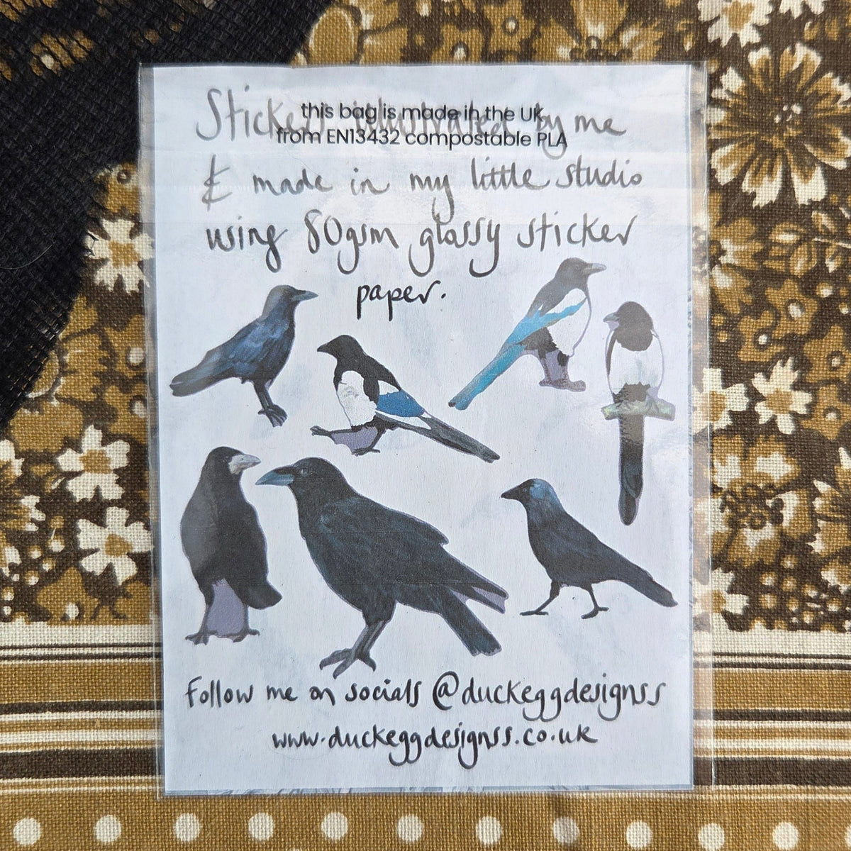 The back of the sticker set showing illustrations of the different corvid stickers inside. Behind the sticker set is a brown floral patterned fabric.