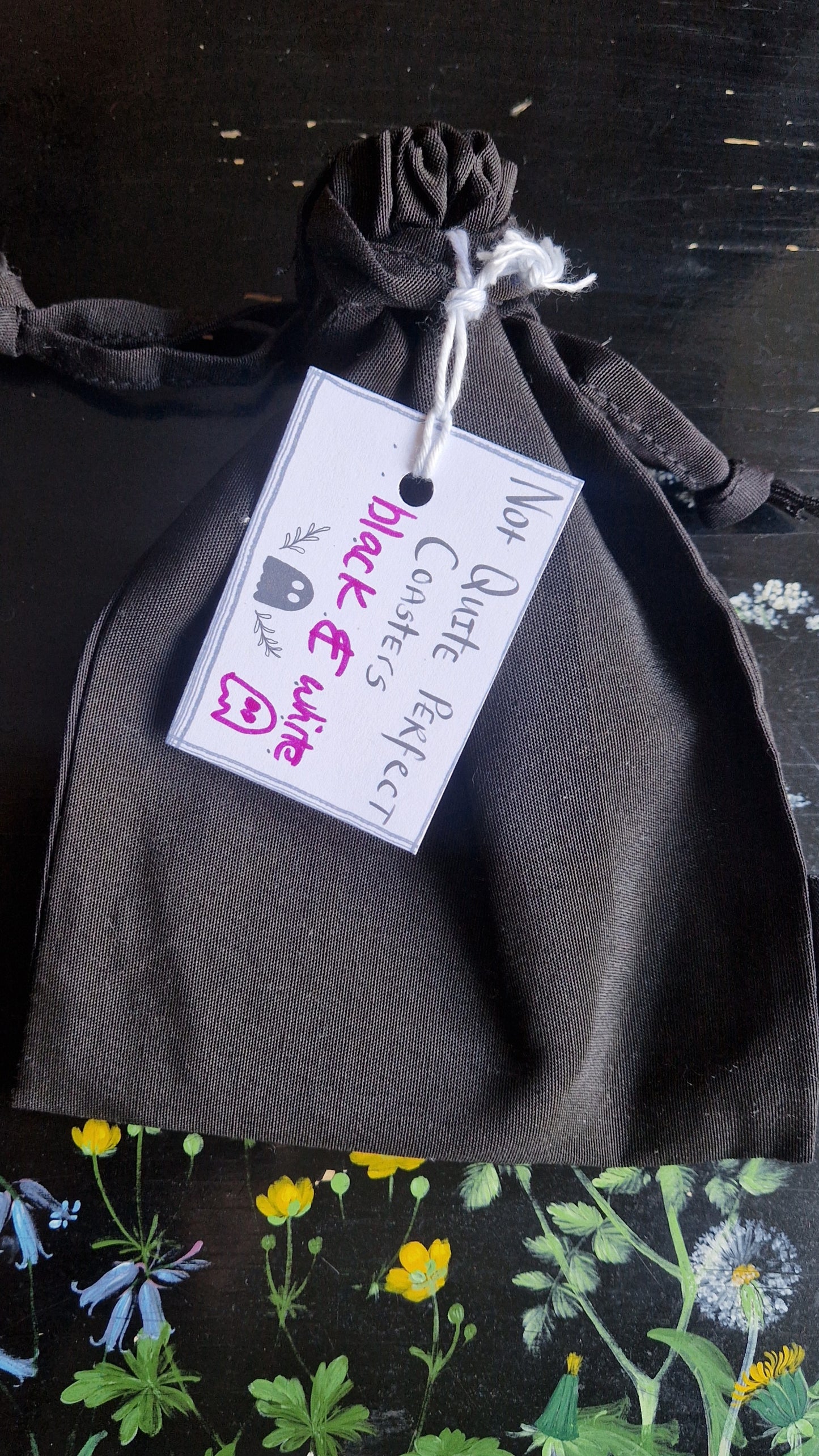 A black fabric drawstring pouch sits on a black table with flowers painted on it. The pouch has a white label that reads 'Not Quite Perfect Coasters' in black writing with the words 'black & white' in purple written on it.