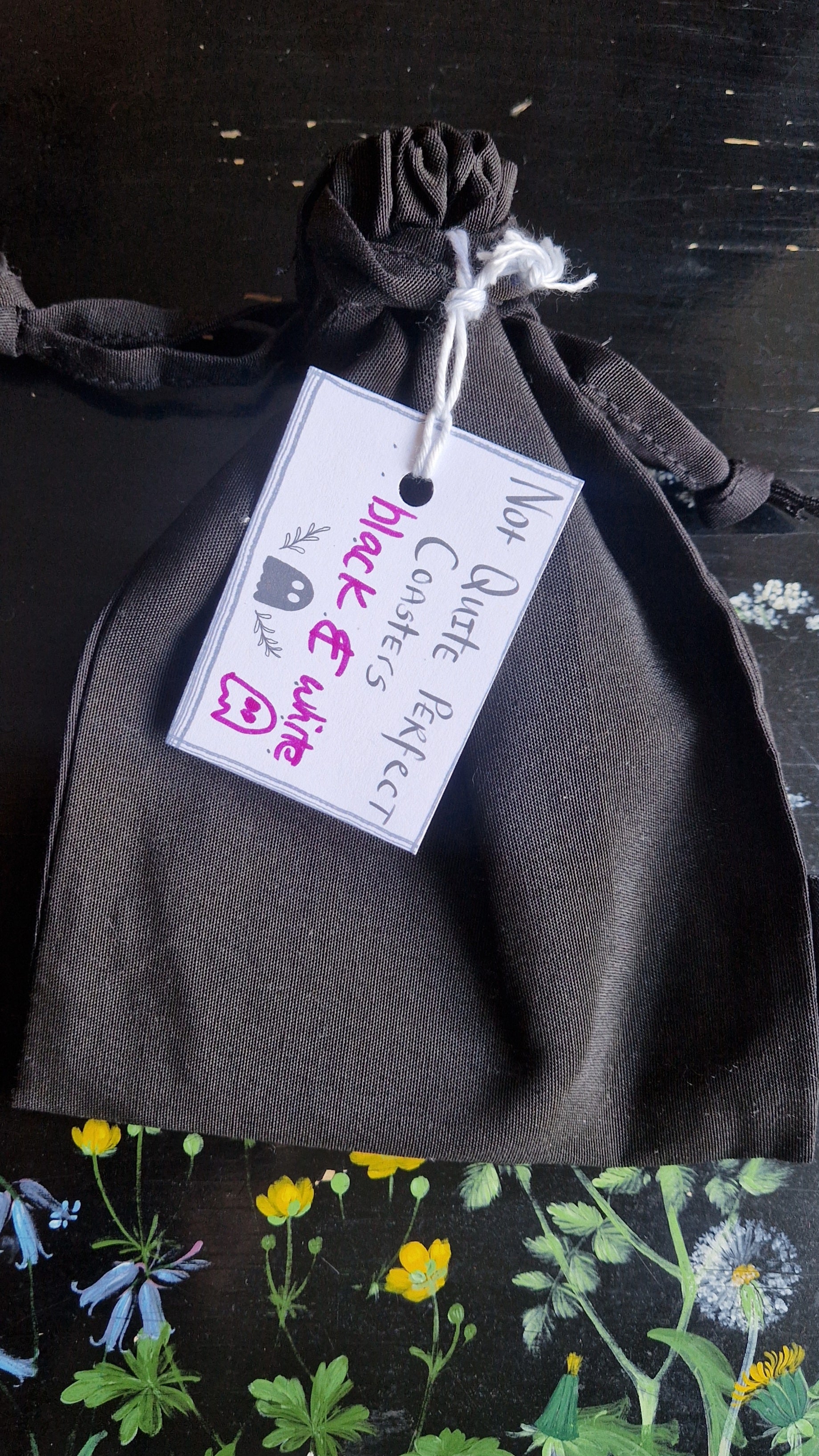 A black fabric drawstring pouch sits on a black table with flowers painted on it. The pouch has a white label that reads 'Not Quite Perfect Coasters' in black writing with the words 'black & white' in purple written on it.