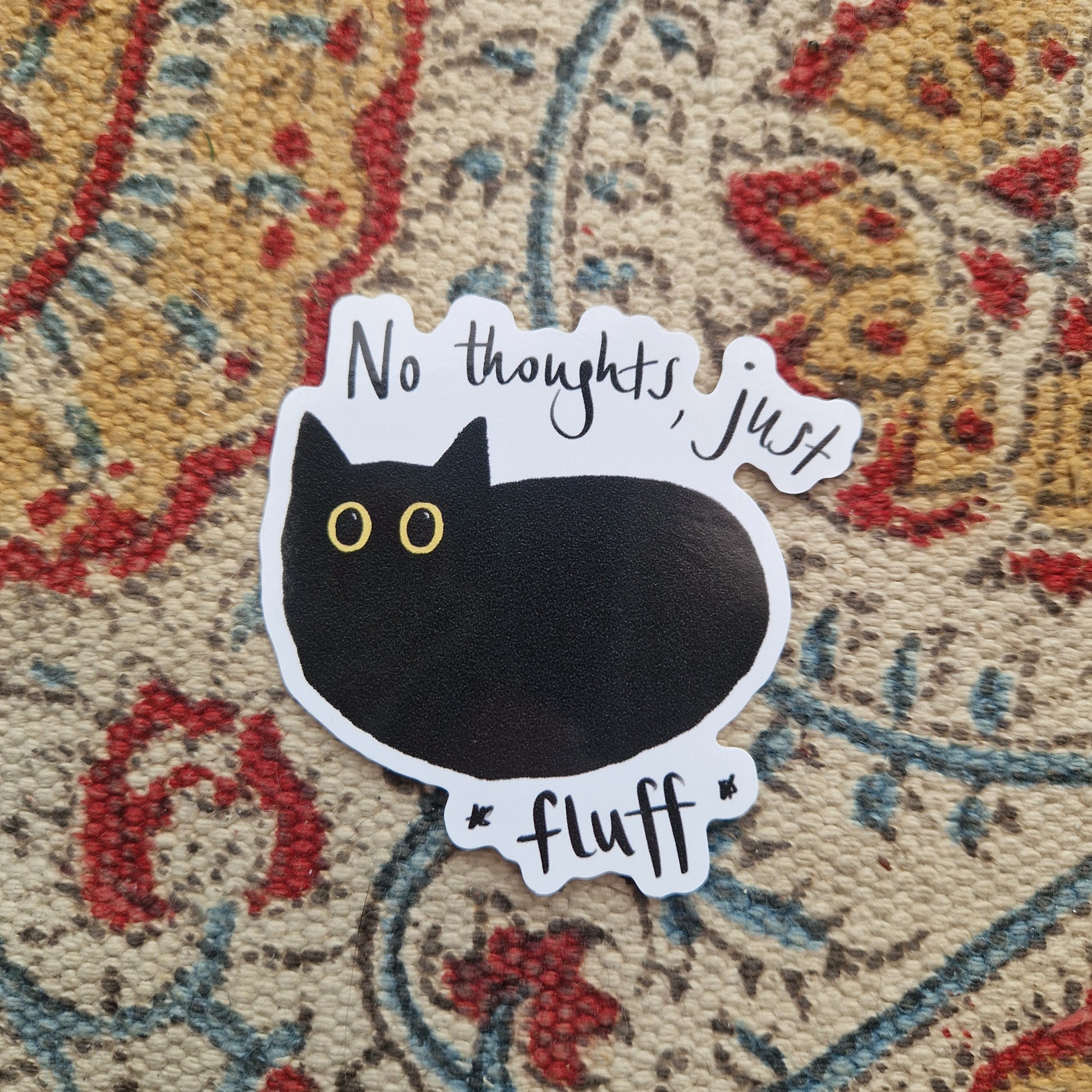 A sticker featuring a simple black cat illustraton with large yellow eyes and the words 'No thoughts, just fluff' written around the cat. Behind the sticker is a patterned fabric background.