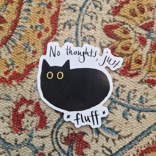 A sticker featuring a simple black cat illustraton with large yellow eyes and the words 'No thoughts, just fluff' written around the cat. Behind the sticker is a patterned fabric background.