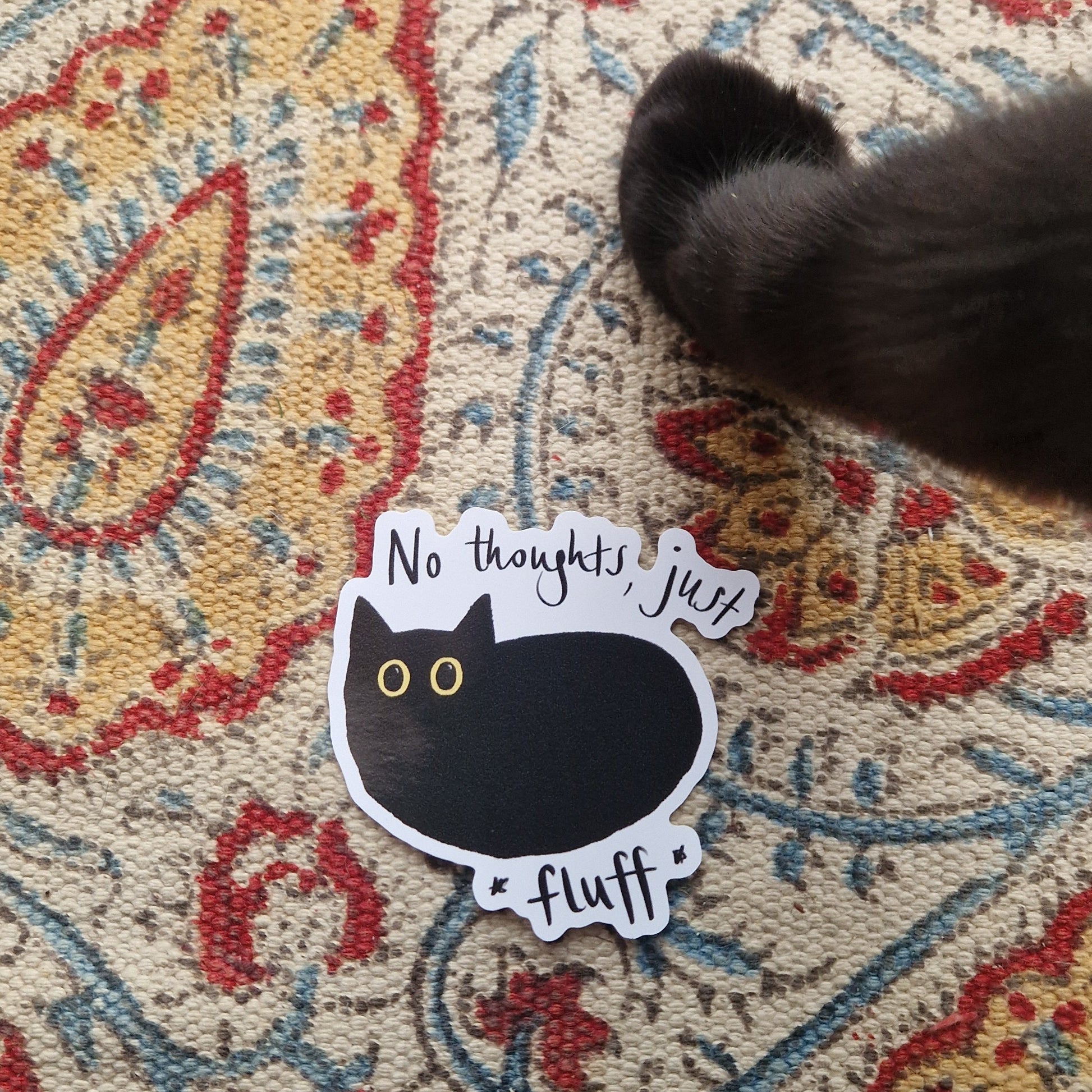 A sticker of a black cat with big yellow eyes and the words 'Not thoughts, just fluff in black handwriting around  it. The sticker sits on a patterned fabric background with a black cat foot just above it (belonging to the cat that inspired it).