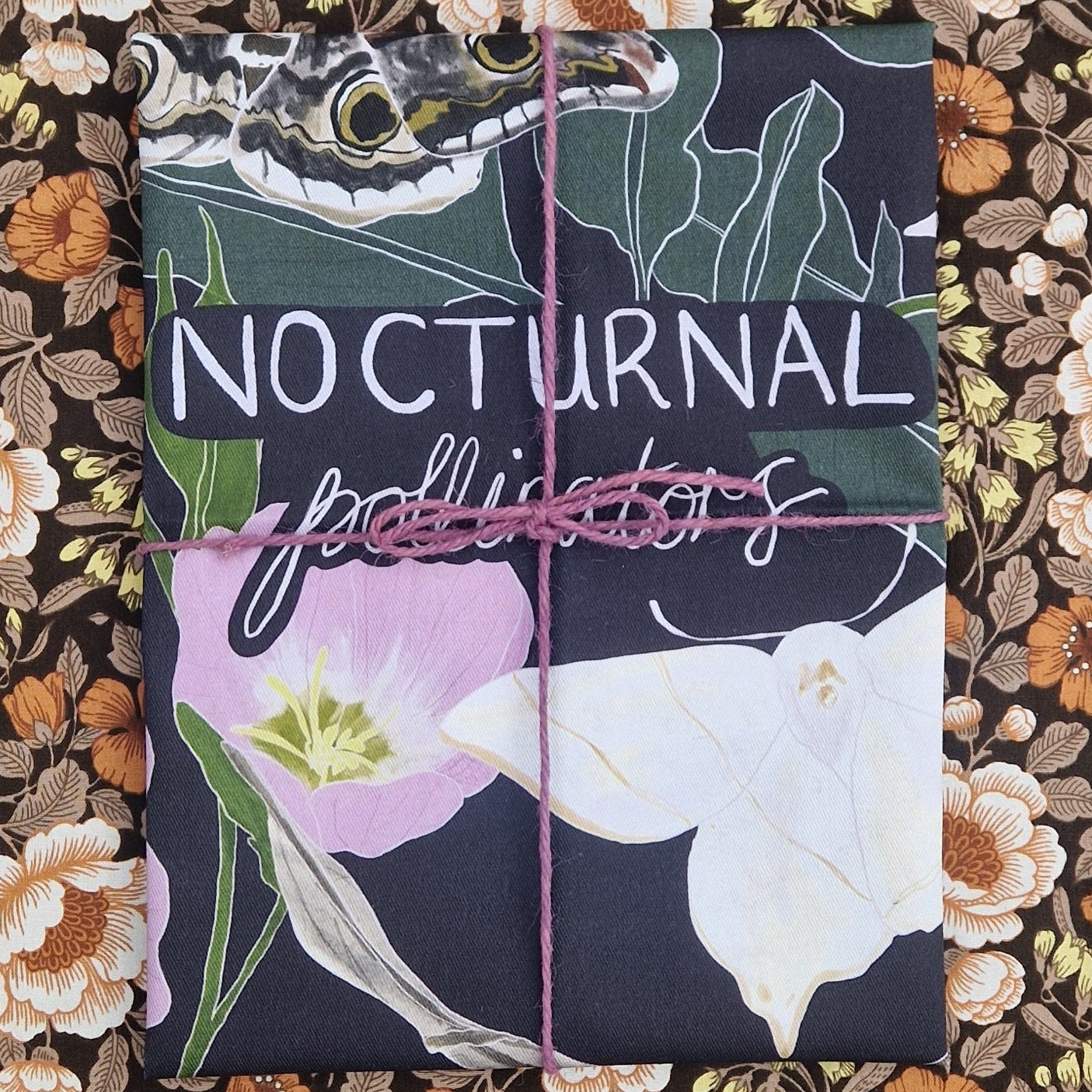 A dark blue folded teatowel wrapped in purple twine sits in front of a floral patterned fabric. The tea towel has the words 'Nocturnal Pollinators' across the middle and two moths as well as a pink flower and green leaves are visible.