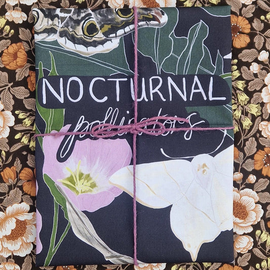 A dark blue folded teatowel wrapped in purple twine sits in front of a floral patterned fabric. The tea towel has the words 'Nocturnal Pollinators' across the middle and two moths as well as a pink flower and green leaves are visible.
