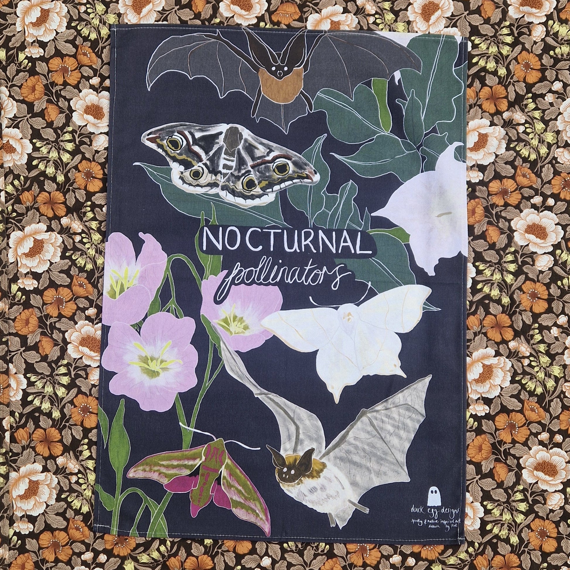A dark blue teatowel lies on top of a patterned floral fabric. The teatowel has the words 'NOCTURNAL pollinators' in white handwriting across the middle surrounded by a designs of night flowering jasmine, bats and moths.