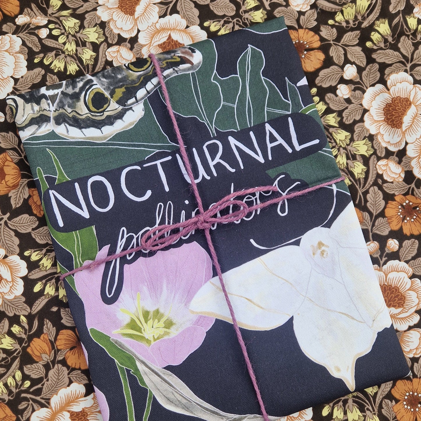 A dark blue folded teatowel wrapped in purple twine sits in front of a floral patterned fabric. The tea towel has the words 'Nocturnal Pollinators' across the middle and two moths as well as a pink flower and green leaves are visible.
