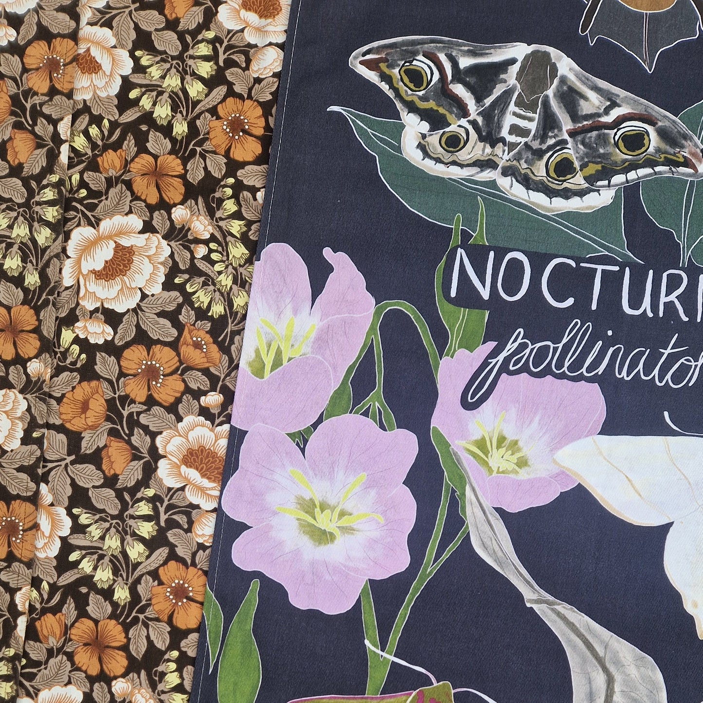 A close up of a dark blue teatowel featuring night flowering pink flowers, moths and part of a bat in view as well as part of the words 'NOCTURNAL pollinators'. To the left of the teatowel is a floral patterned fabric.