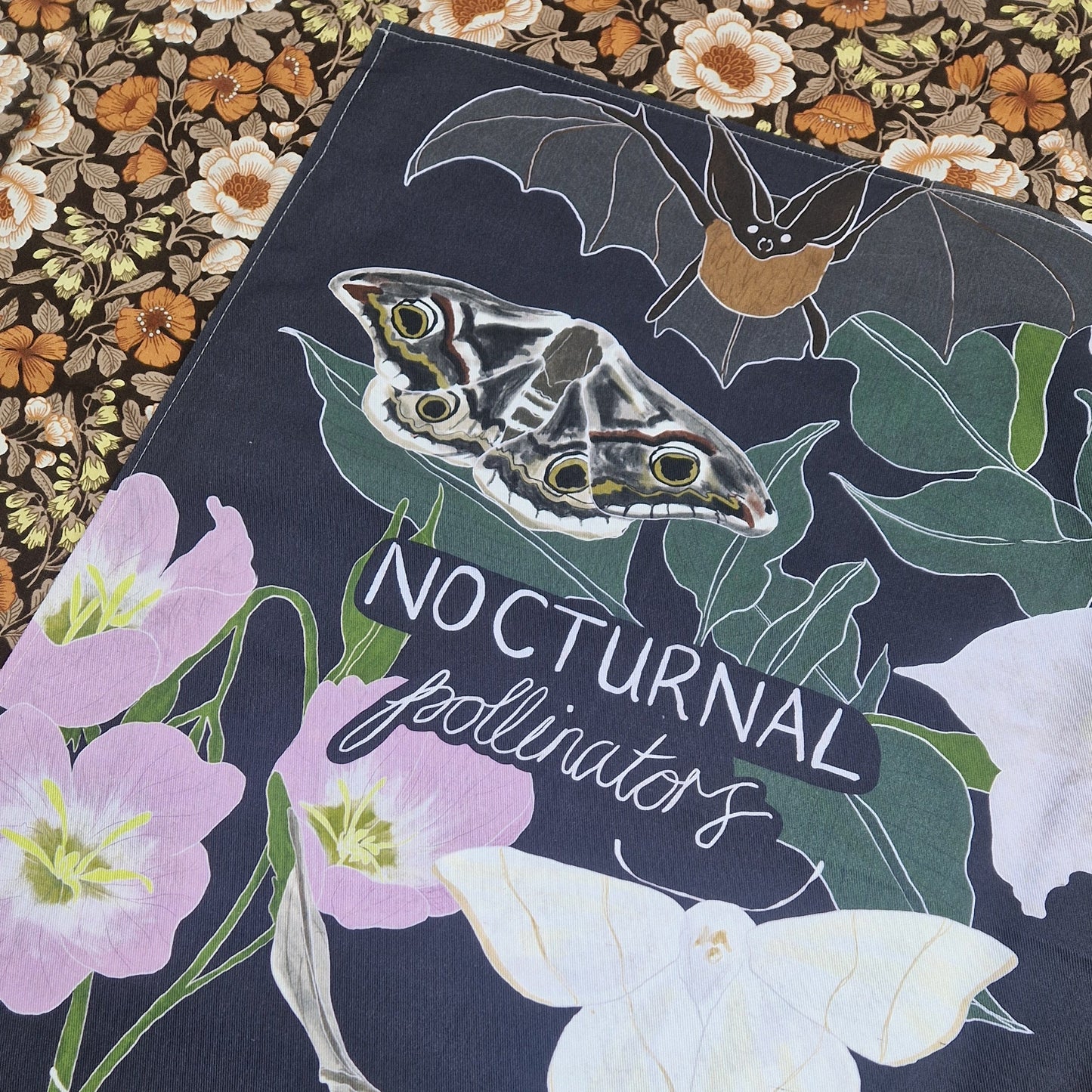 A dark blue teatowel lies on top of a patterned floral fabric. The teatowel has the words 'NOCTURNAL pollinators' in white handwriting across the middle surrounded by a designs of night flowering jasmine, bats and moths.