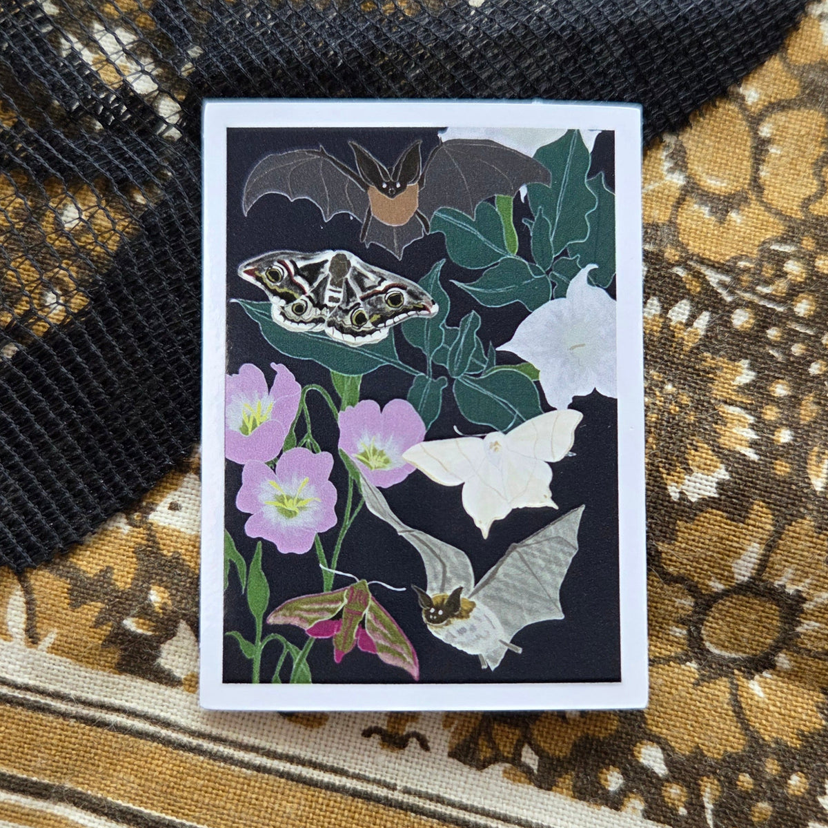 A rectangular sticker with a white border and a dark blue background with nocturnal moths, bats and plants sits in front of a brown patterned fabric with a black lace overlay.