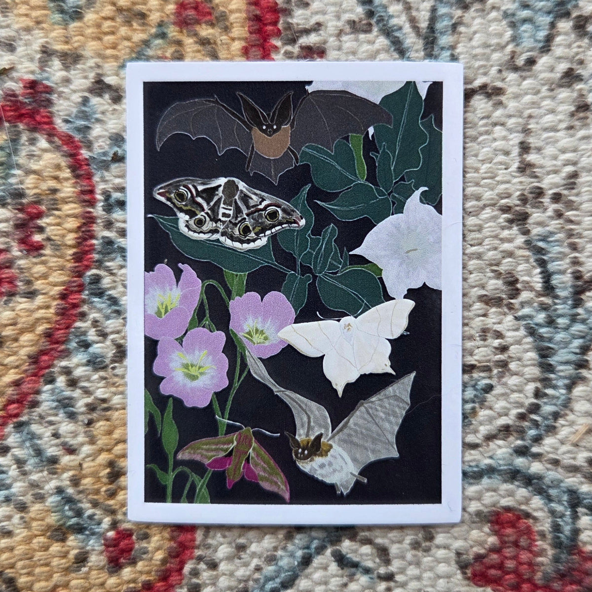 A rectangular sticker with a white border and a dark blue background with nocturnal moths, bats and plants sits in front of a colourful patterned fabric.