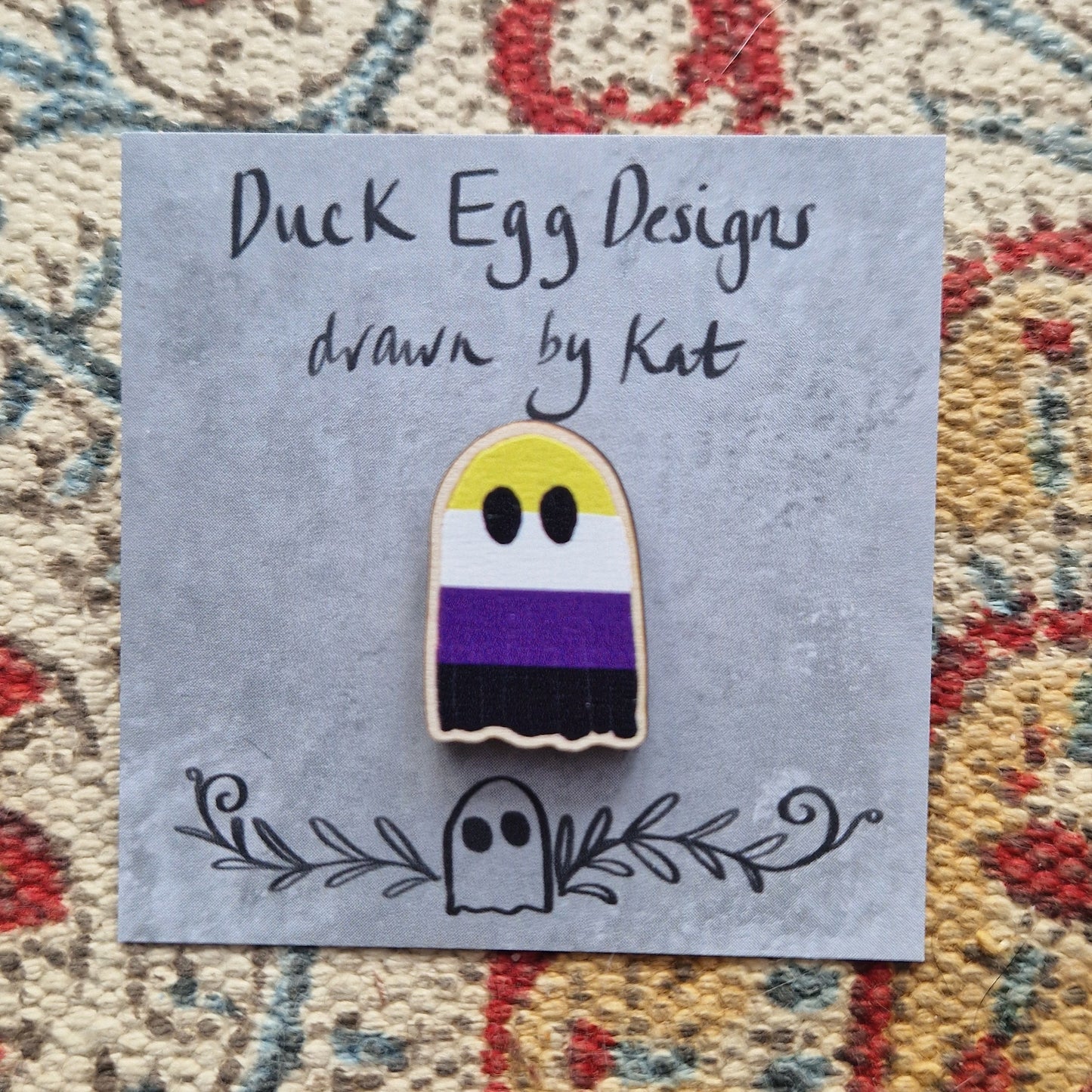 A ghost pin badge featuring the striped design of the non-binary flag (yellow, white, purple and black) sits on a grey backing card with the words 'Duck Egg Designs drawn by Kat' in black handwriting across the top. Behind the backing card is a patterned fabric background.