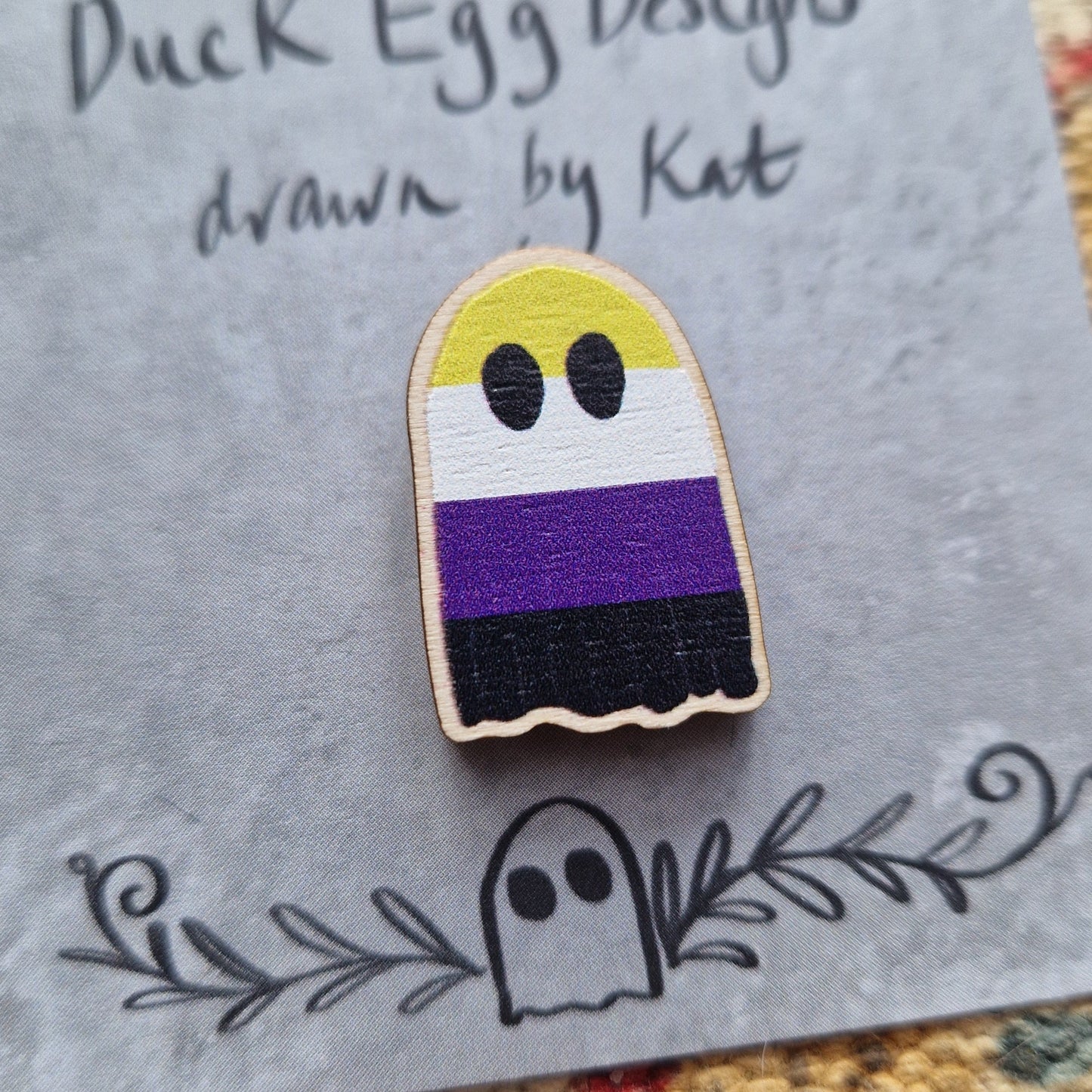 A ghost pin badge featuring the striped design of the non-binary flag (yellow, white, purple and black) sits on a grey backing card with the words 'Duck Egg Designs drawn by Kat' in black handwriting across the top. Behind the backing card is a patterned fabric background.