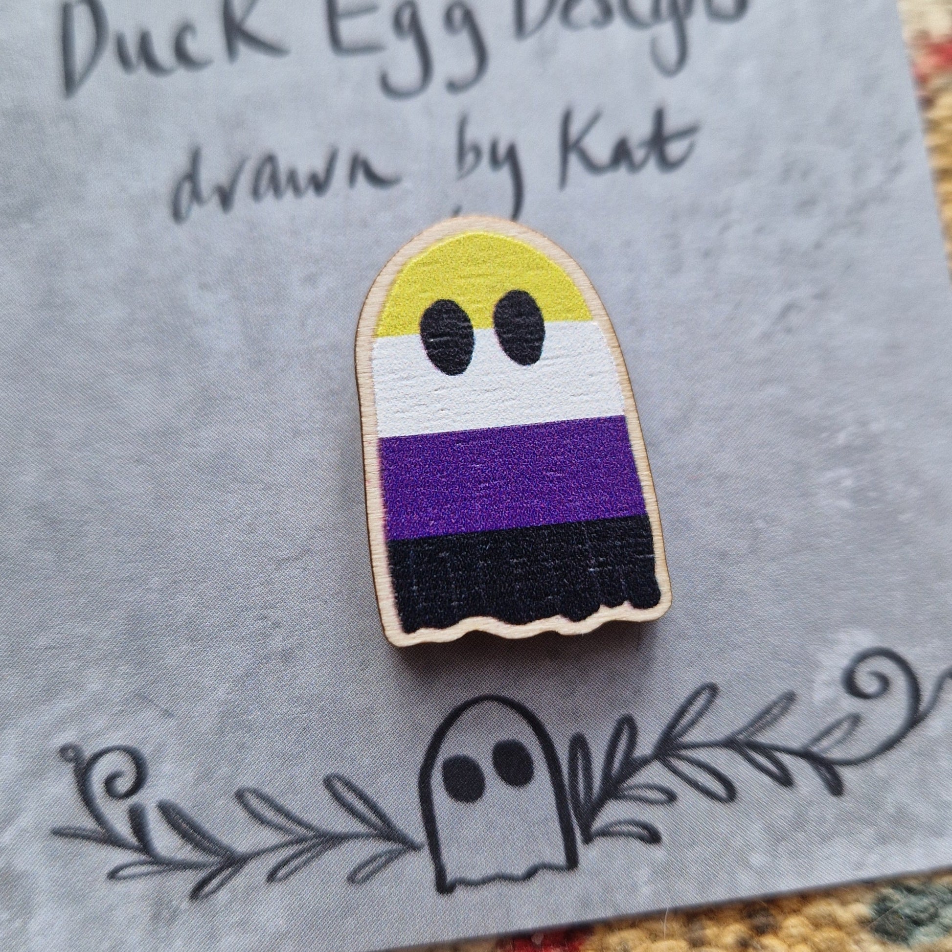 A ghost pin badge featuring the striped design of the non-binary flag (yellow, white, purple and black) sits on a grey backing card with the words 'Duck Egg Designs drawn by Kat' in black handwriting across the top. Behind the backing card is a patterned fabric background.