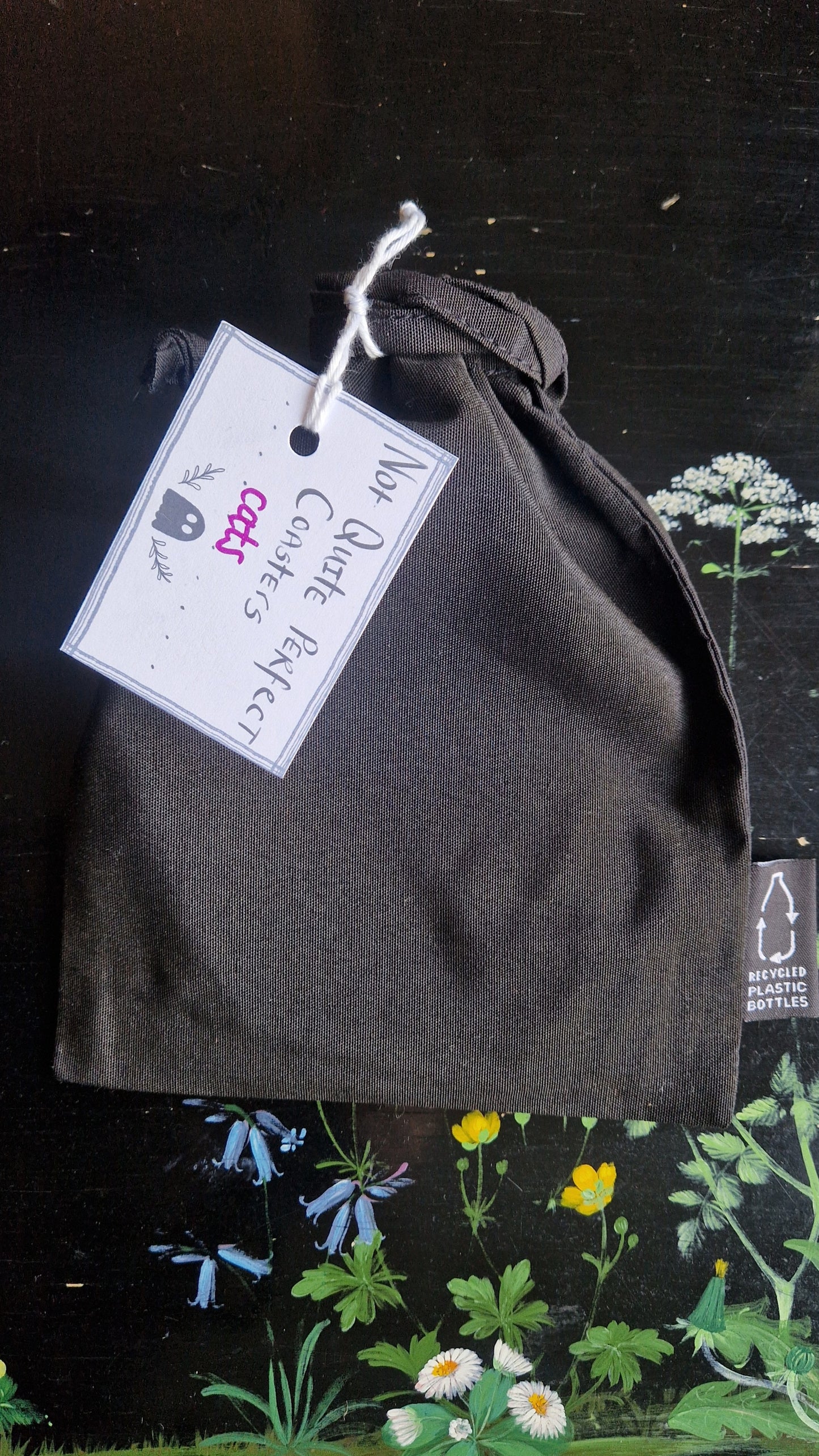 A black fabric drawstring pouch sits on a black table with flowers painted on it. The pouch has a white label that reads 'Not Quite Perfect Coasters' in black writing with the word 'cats' in purple written on it.