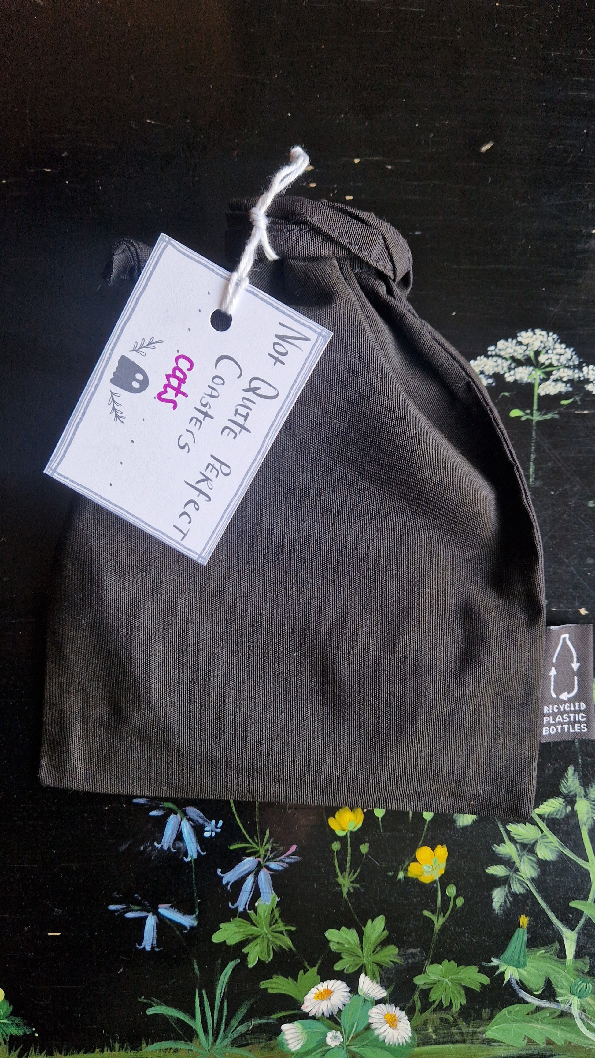A black fabric drawstring pouch sits on a black table with flowers painted on it. The pouch has a white label that reads 'Not Quite Perfect Coasters' in black writing with the word 'cats' in purple written on it.