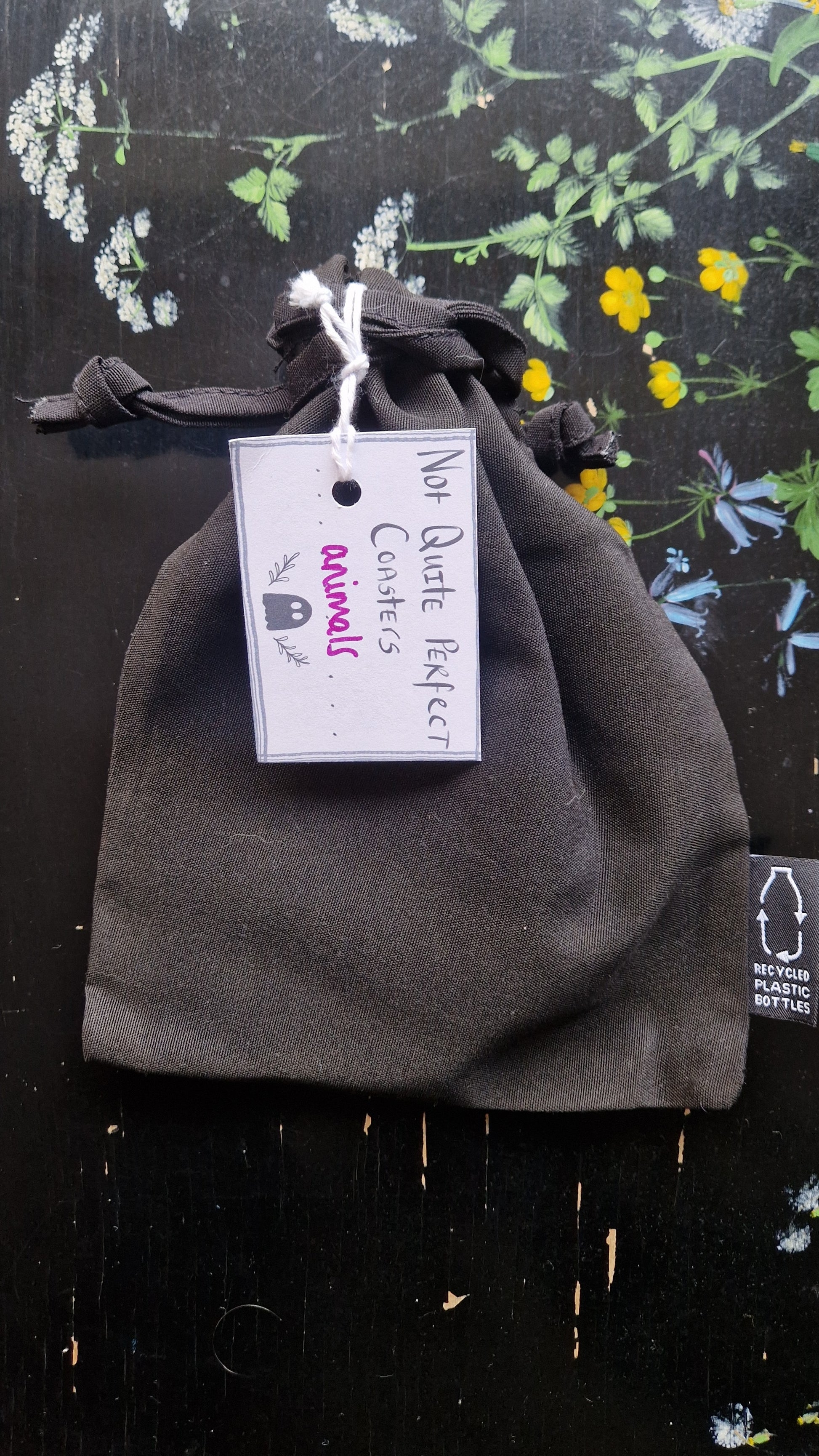 A black fabric drawstring pouch sits on a black table with flowers painted on it. The pouch has a white label that reads 'Not Quite Perfect Coasters' in black writing with the word 'animals' in purple written on it.