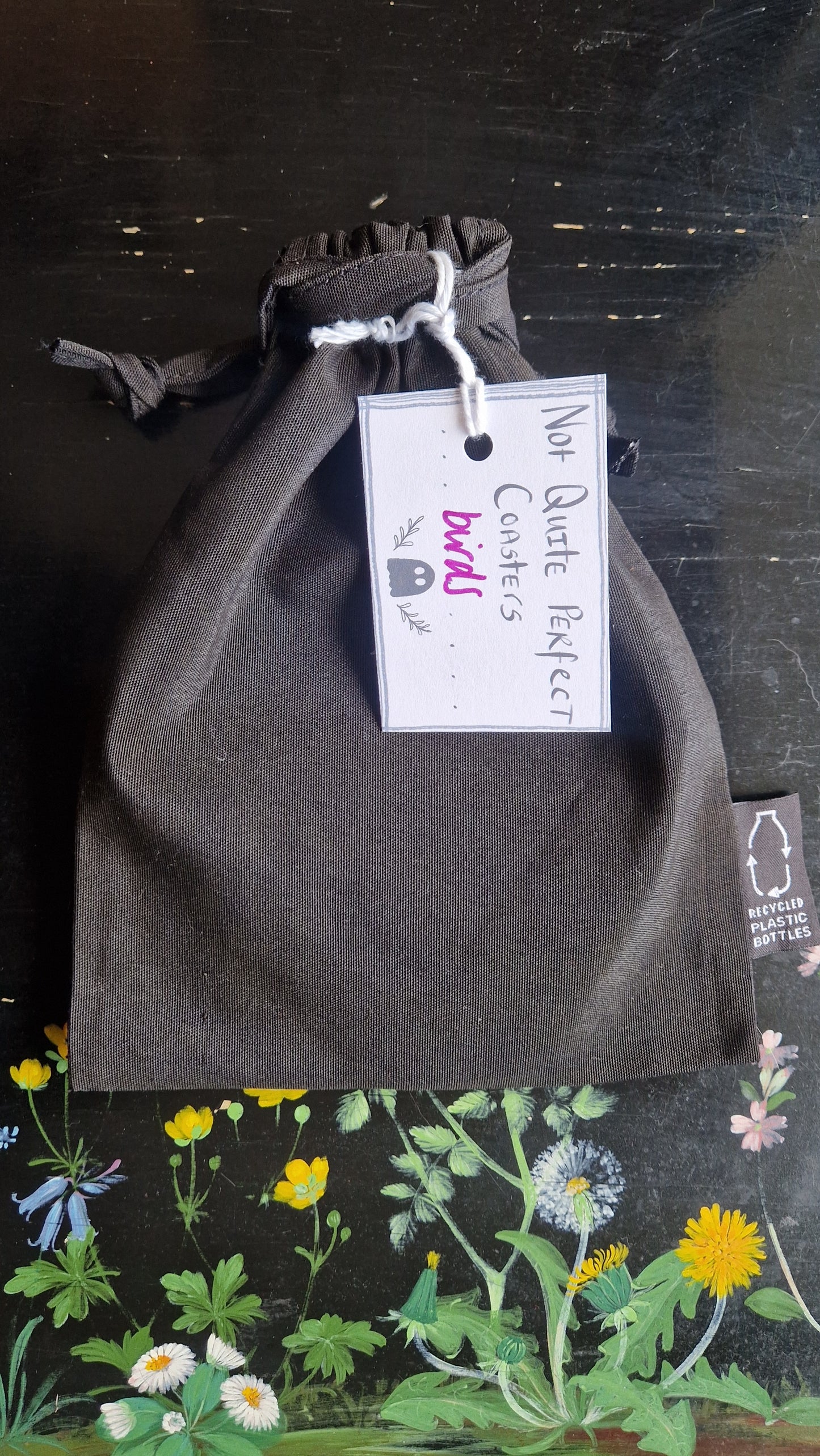 A black fabric drawstring pouch sits on a black table with flowers painted on it. The pouch has a white label that reads 'Not Quite Perfect Coasters' in black writing with the word 'birds' in purple written on it.