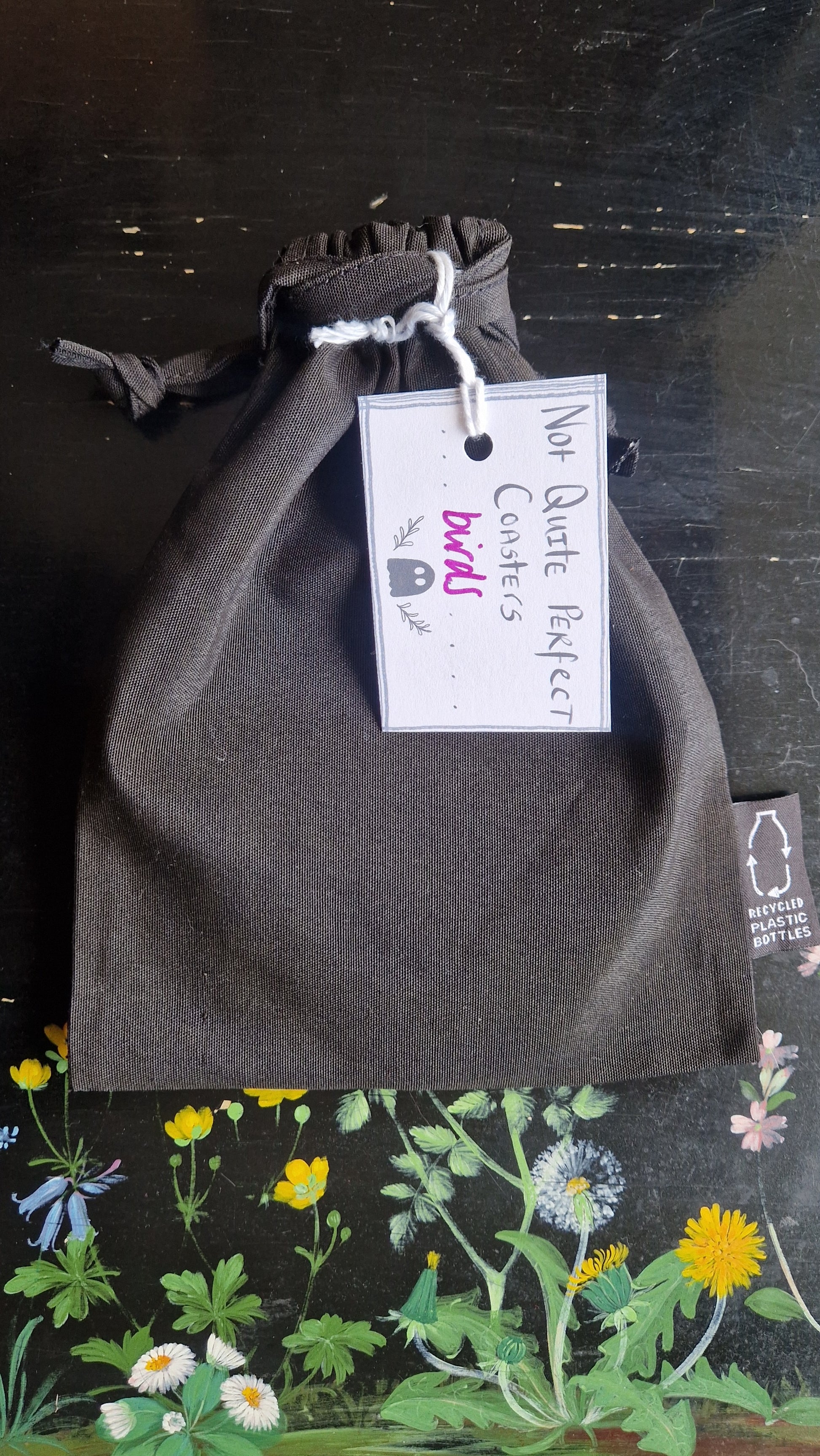 A black fabric drawstring pouch sits on a black table with flowers painted on it. The pouch has a white label that reads 'Not Quite Perfect Coasters' in black writing with the word 'birds' in purple written on it.