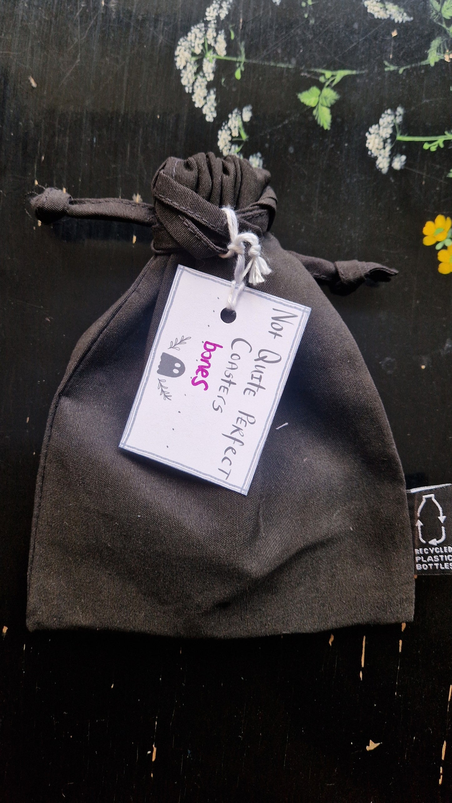 A black fabric drawstring pouch sits on a black table with flowers painted on it. The pouch has a white label that reads 'Not Quite Perfect Coasters' in black writing with the word 'bones' in purple written on it.