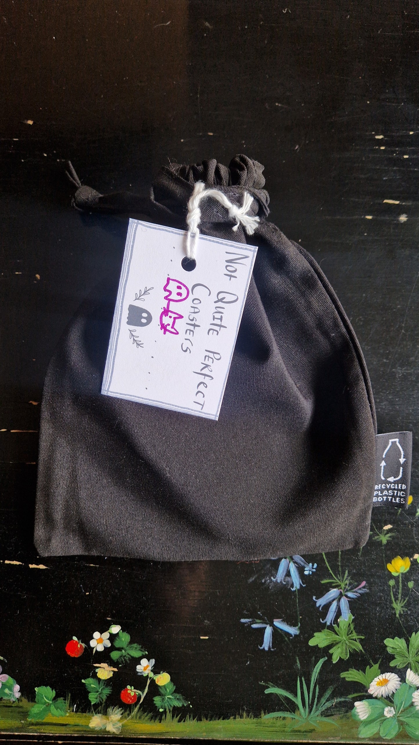 A black fabric drawstring pouch sits on a black table with flowers painted on it. The pouch has a white label that reads 'Not Quite Perfect Coasters' in black writing with the line drawings of a ghost and a cat head in purple written on it.