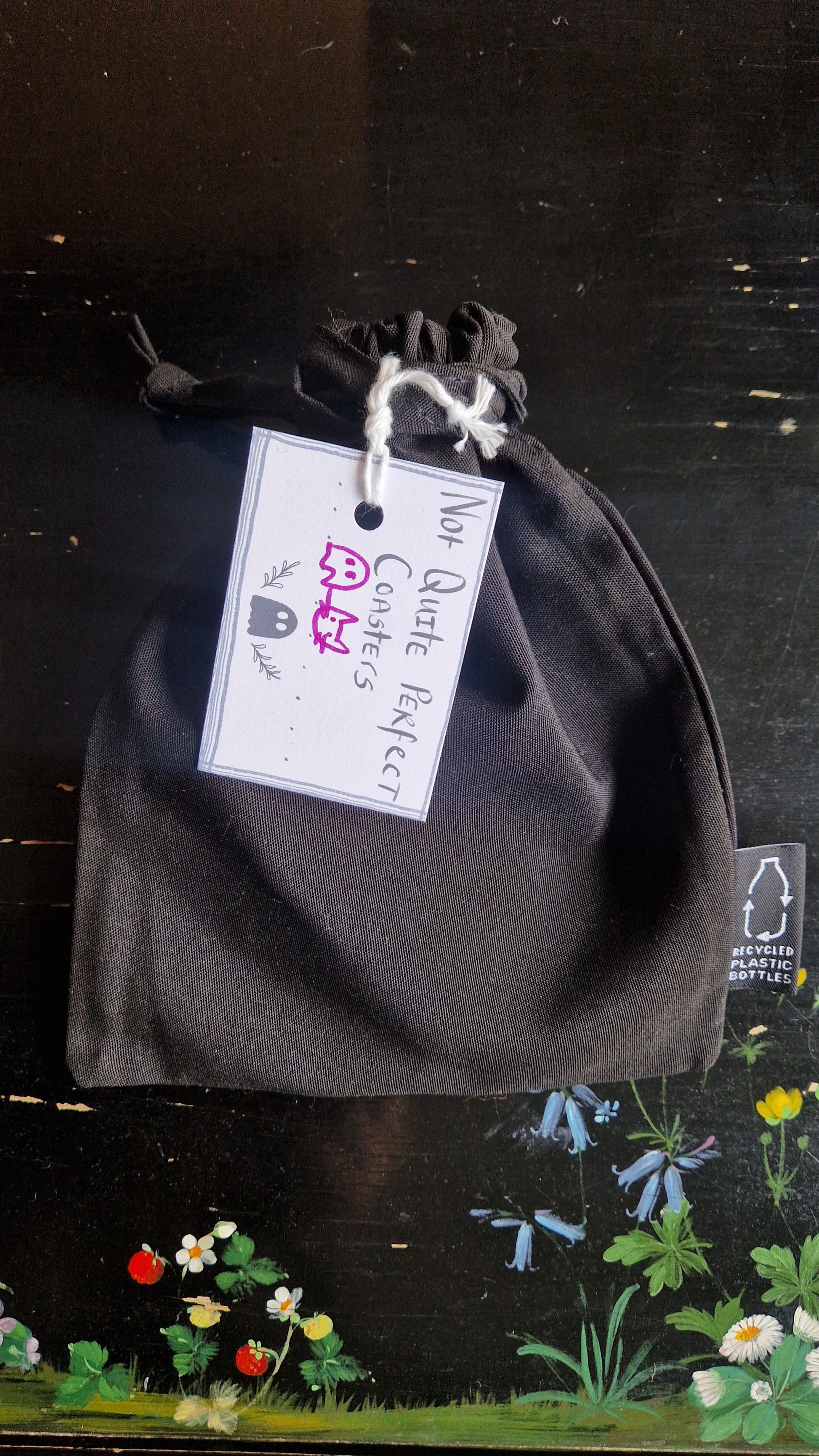 A black fabric drawstring pouch sits on a black table with flowers painted on it. The pouch has a white label that reads 'Not Quite Perfect Coasters' in black writing with the line drawings of a ghost and a cat head in purple written on it.
