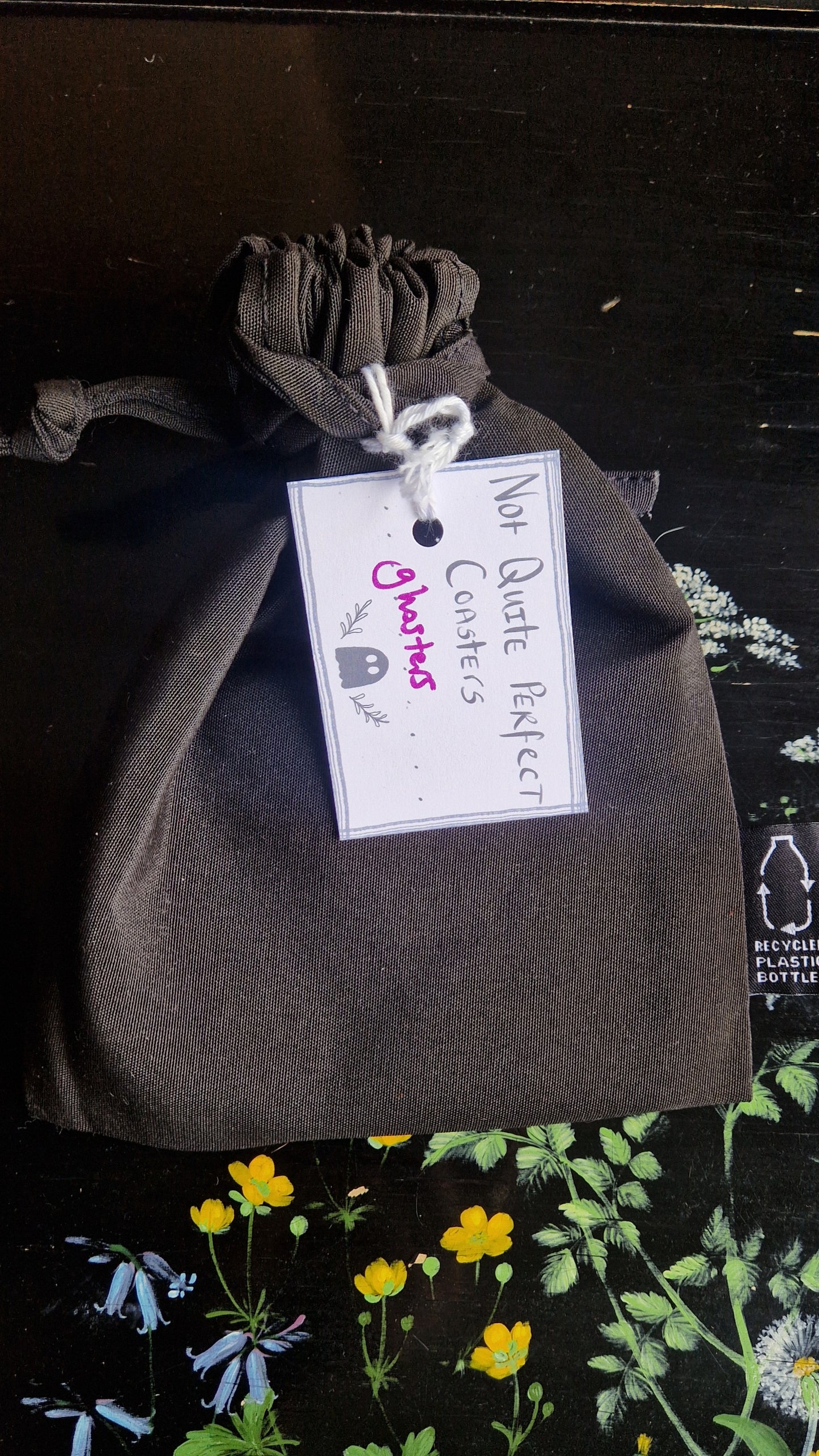 A black fabric drawstring pouch sits on a black table with flowers painted on it. The pouch has a white label that reads 'Not Quite Perfect Coasters' in black writing with the word 'ghosters' in purple written on it.