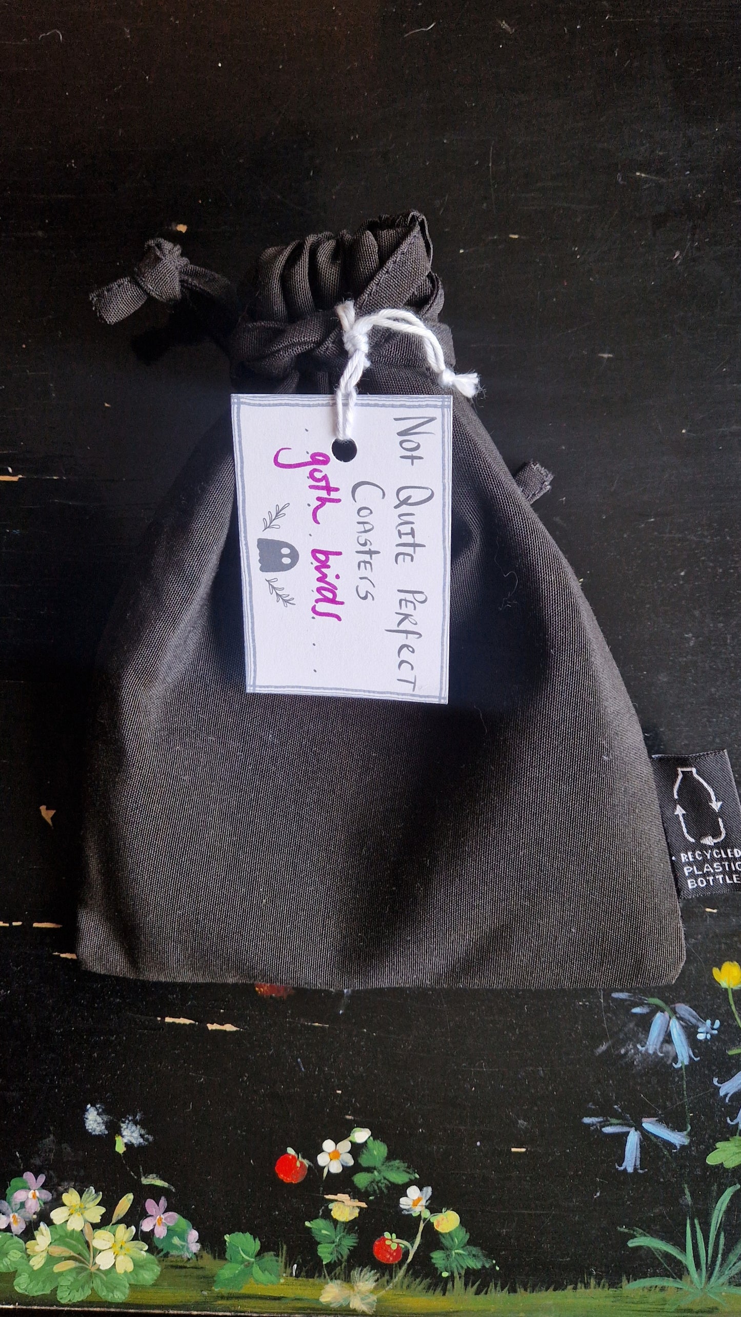 A black fabric drawstring pouch sits on a black table with flowers painted on it. The pouch has a white label that reads 'Not Quite Perfect Coasters' in black writing with the words 'goth birds' in purple written on it.