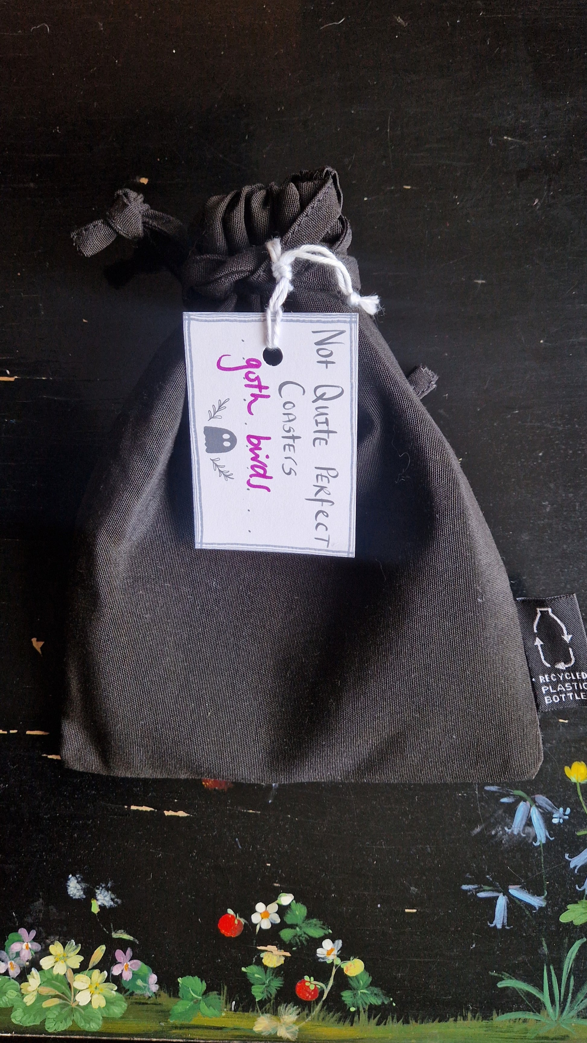 A black fabric drawstring pouch sits on a black table with flowers painted on it. The pouch has a white label that reads 'Not Quite Perfect Coasters' in black writing with the words 'goth birds' in purple written on it.