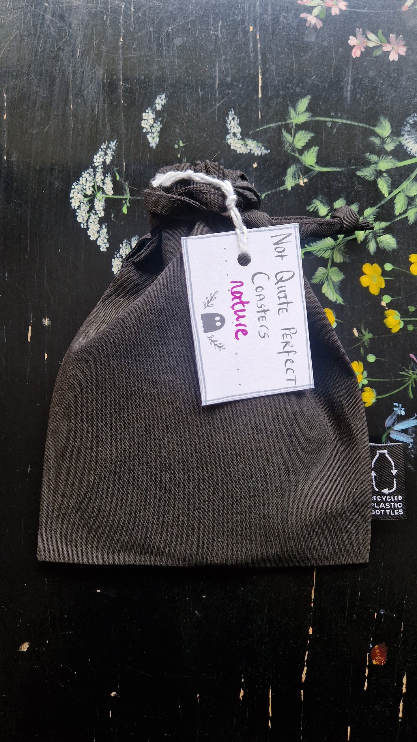 A black fabric drawstring pouch sits on a black table with flowers painted on it. The pouch has a white label that reads 'Not Quite Perfect Coasters' in black writing with the word 'nature' in purple written on it.
