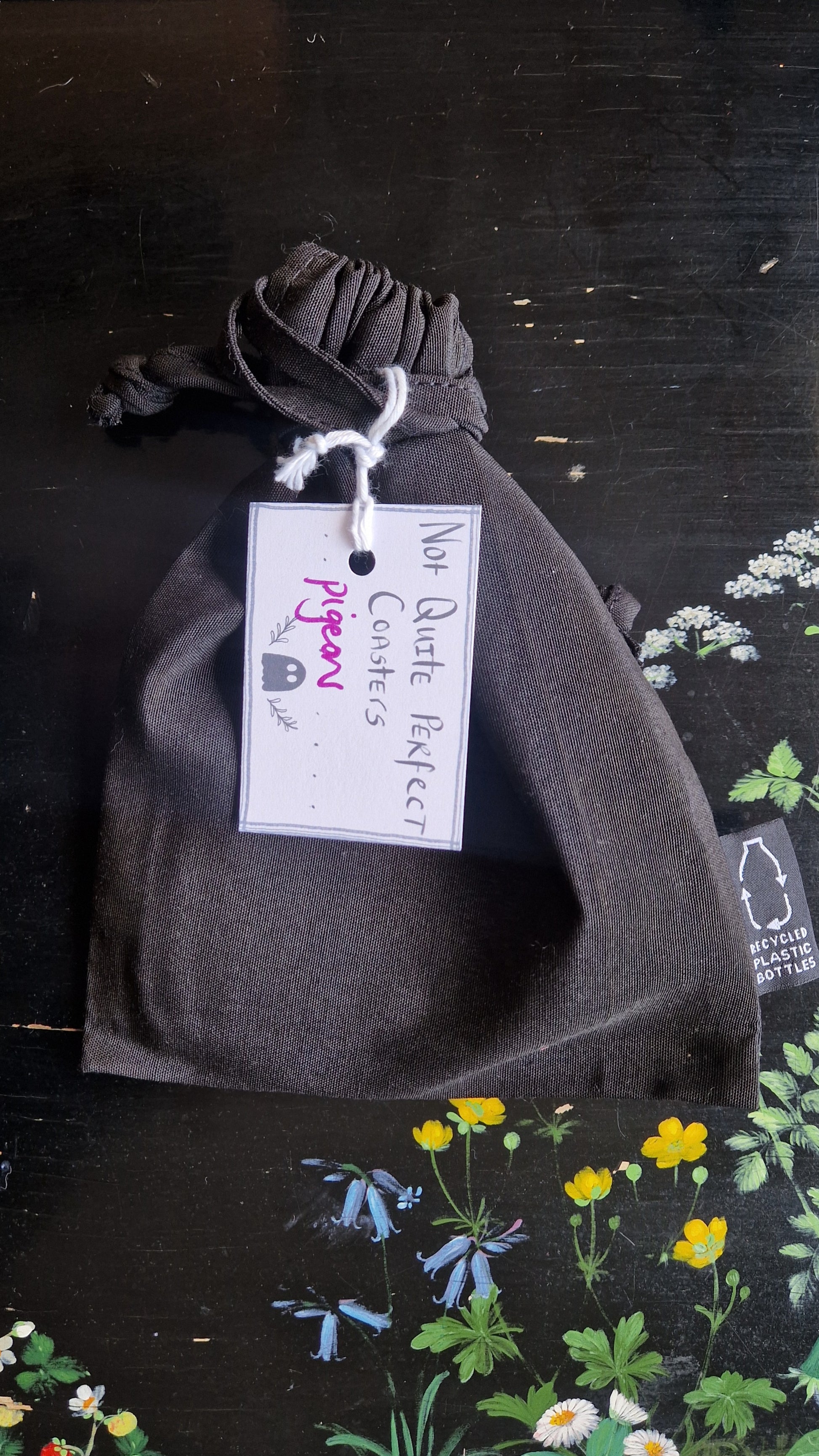 A black fabric drawstring pouch sits on a black table with flowers painted on it. The pouch has a white label that reads 'Not Quite Perfect Coasters' in black writing with the word 'pigeons' in purple written on it.
