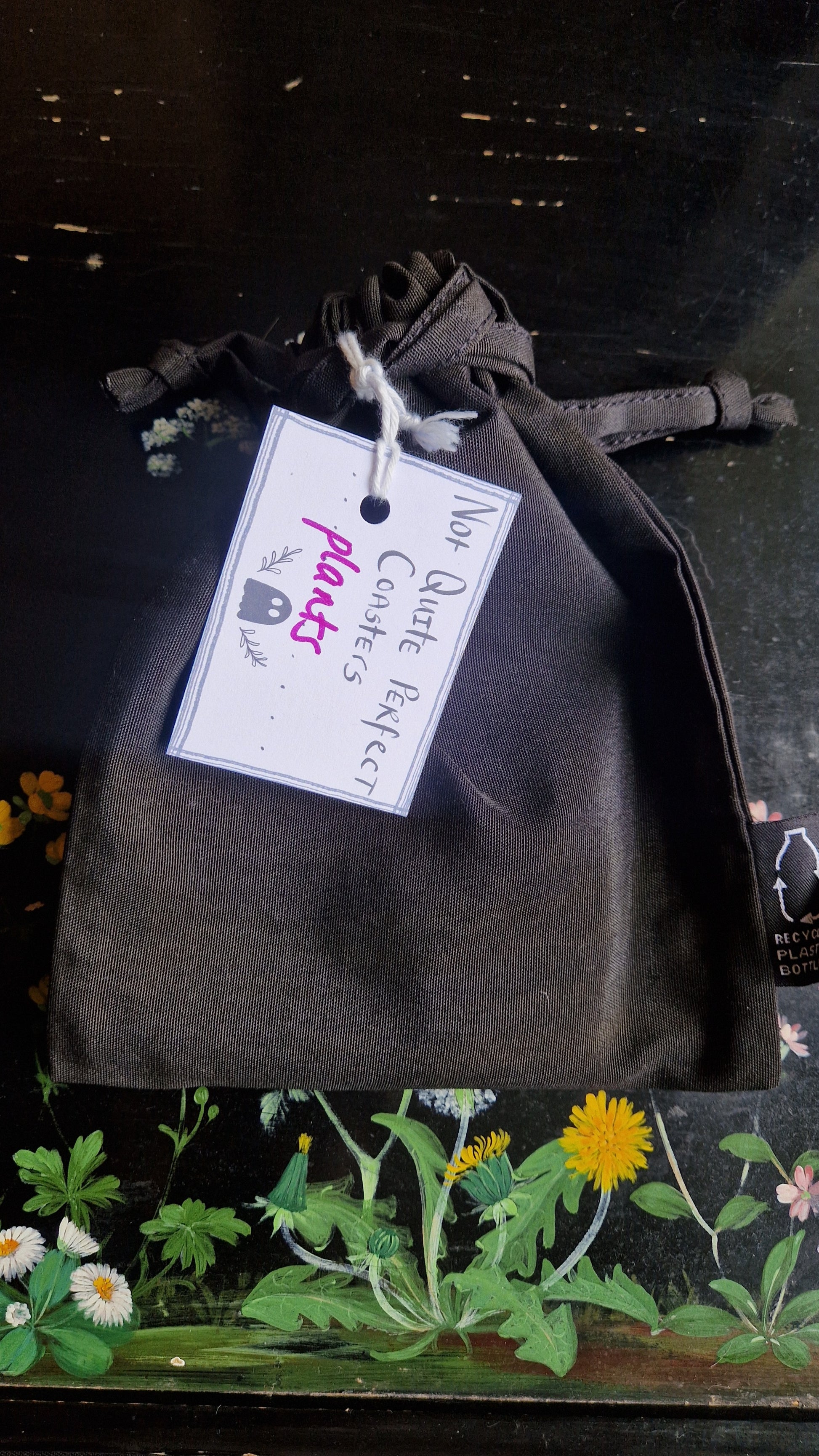 A black fabric drawstring pouch sits on a black table with flowers painted on it. The pouch has a white label that reads 'Not Quite Perfect Coasters' in black writing with the word 'plants' in purple written on it.