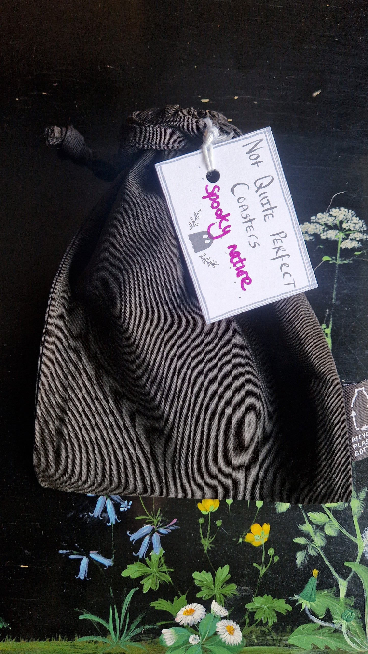 A black fabric drawstring pouch sits on a black table with flowers painted on it. The pouch has a white label that reads 'Not Quite Perfect Coasters' in black writing with the words 'spooky nature' in purple written on it.