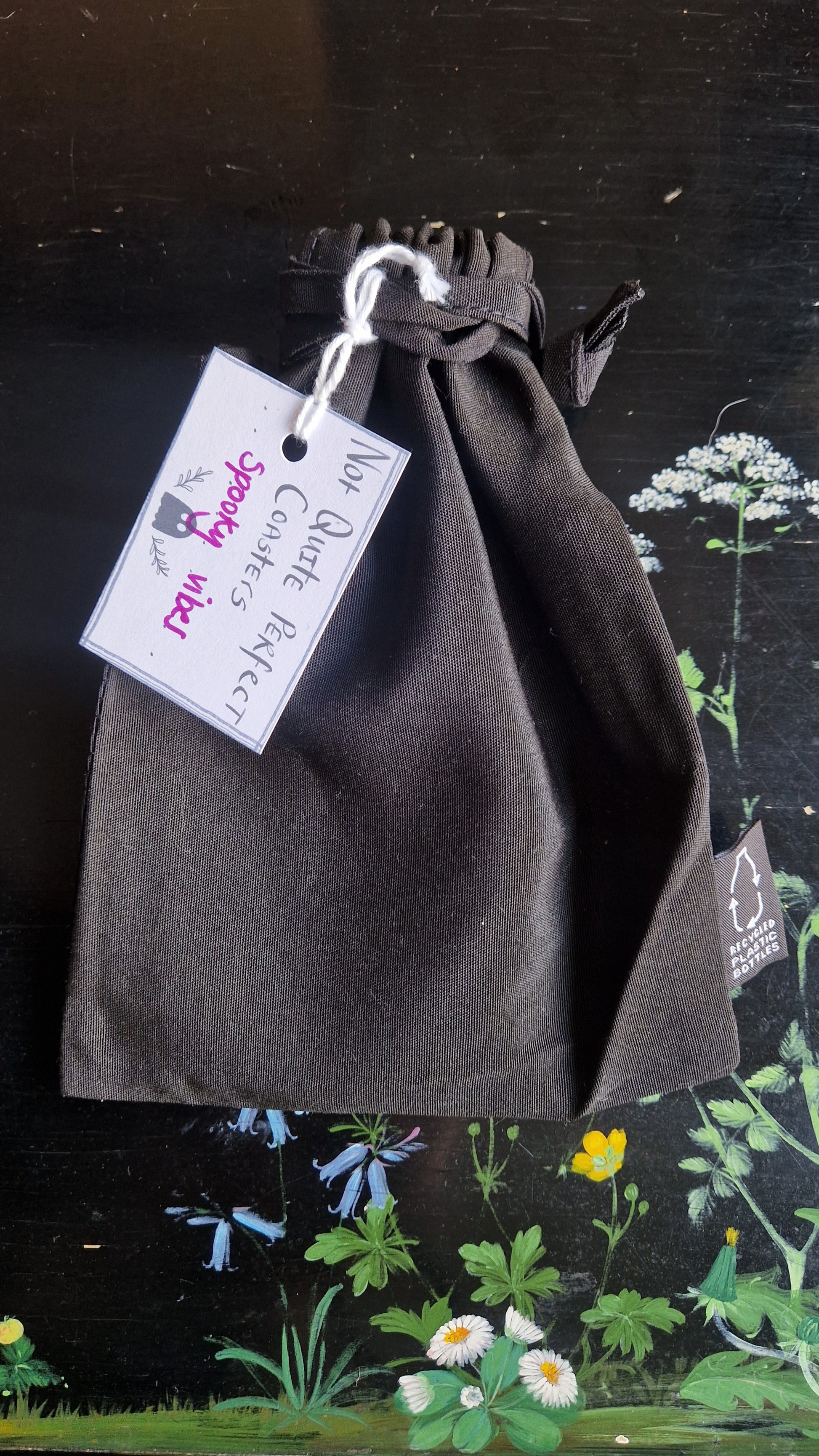 A black fabric drawstring pouch sits on a black table with flowers painted on it. The pouch has a white label that reads 'Not Quite Perfect Coasters' in black writing with the words 'spooky vibes' in purple written on it.