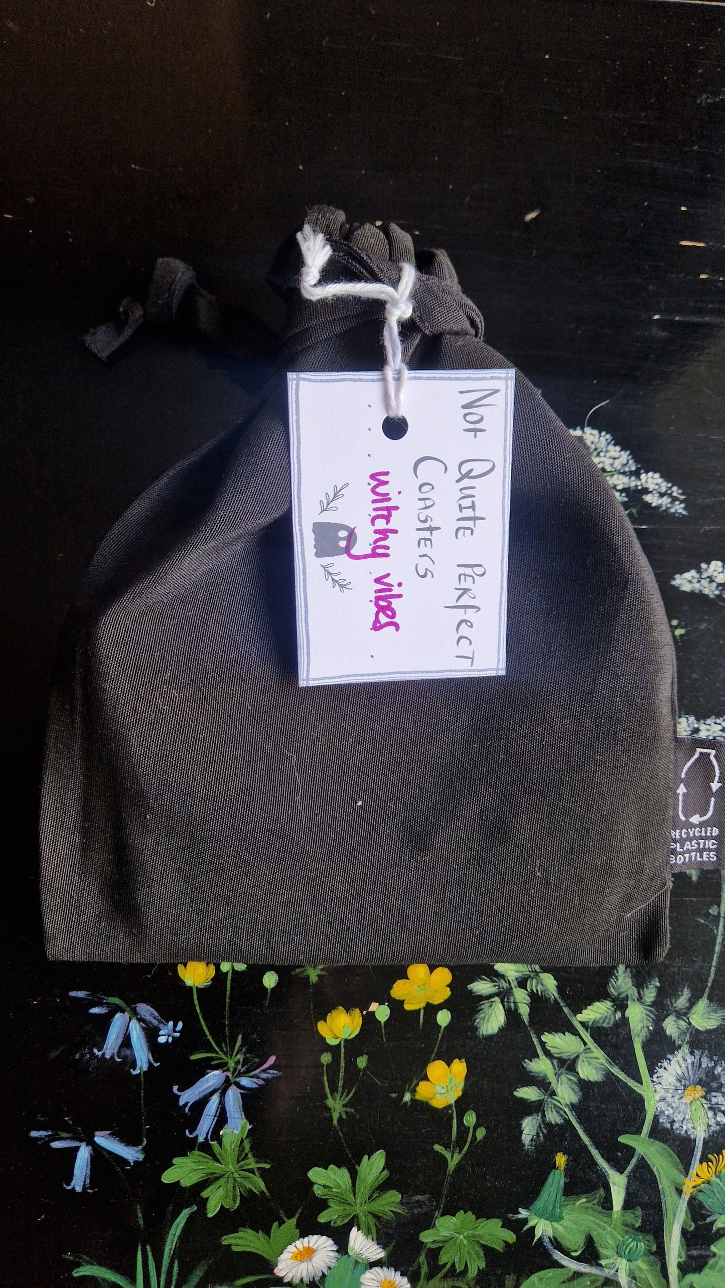 A black fabric drawstring pouch sits on a black table with flowers painted on it. The pouch has a white label that reads 'Not Quite Perfect Coasters' in black writing with the words 'witchy vibes' in purple written on it.
