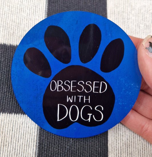 A round blue coaster is held by a white hand in front of a checkered black and white background. The coaster has a black pawprint design with the words 'Obsessed with Dogs' in white handwriting.
