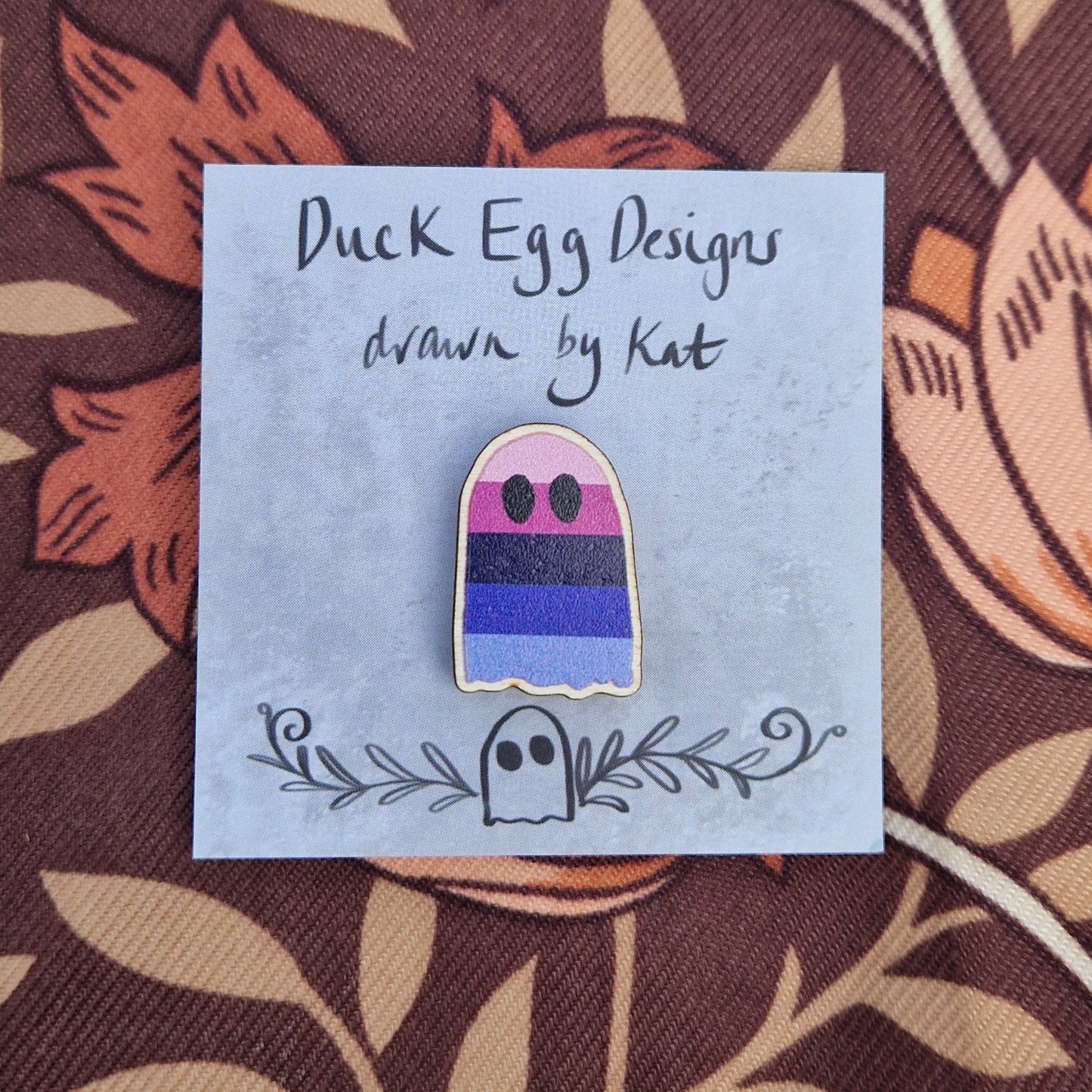 A ghost pin featuring the colourful stripes of the omnisexual flag sits on a grey square backing card with the words 'Duck Egg Designs drawn by Kat' in black across the top. The backing card sits on a brown patterned fabric.