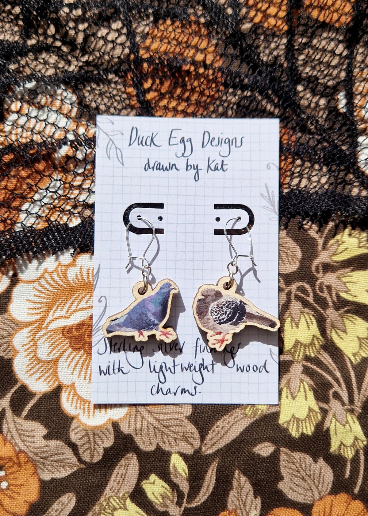 A pair of mismatched pigeon earrings sit on a white backing card with the words 'Duck Egg Designs drawn by Kat' across the top. Behind the backing card is a floral patterned fabric with a black ace overlay. The pigeon on the left is blue, purple and light grey with orange feet while the pigeon on the right is grey, black and white with orange feet and has a chunkier shape.