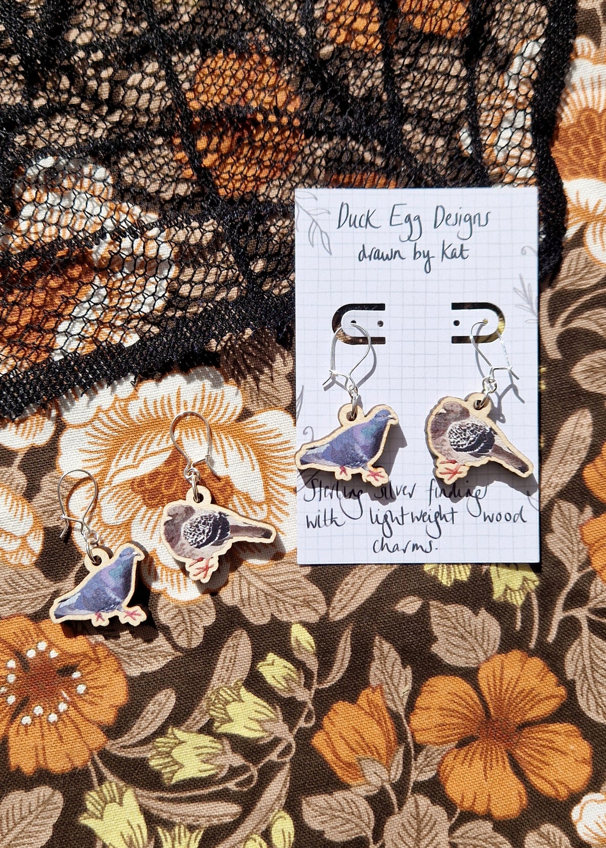 Two pairs of pigeon earrings sit next to each other on a floral fabric background. The right pair of earrings sit on a white backing card with the words 'Duck Egg Designs drawn by Kat' in black handwriting. 