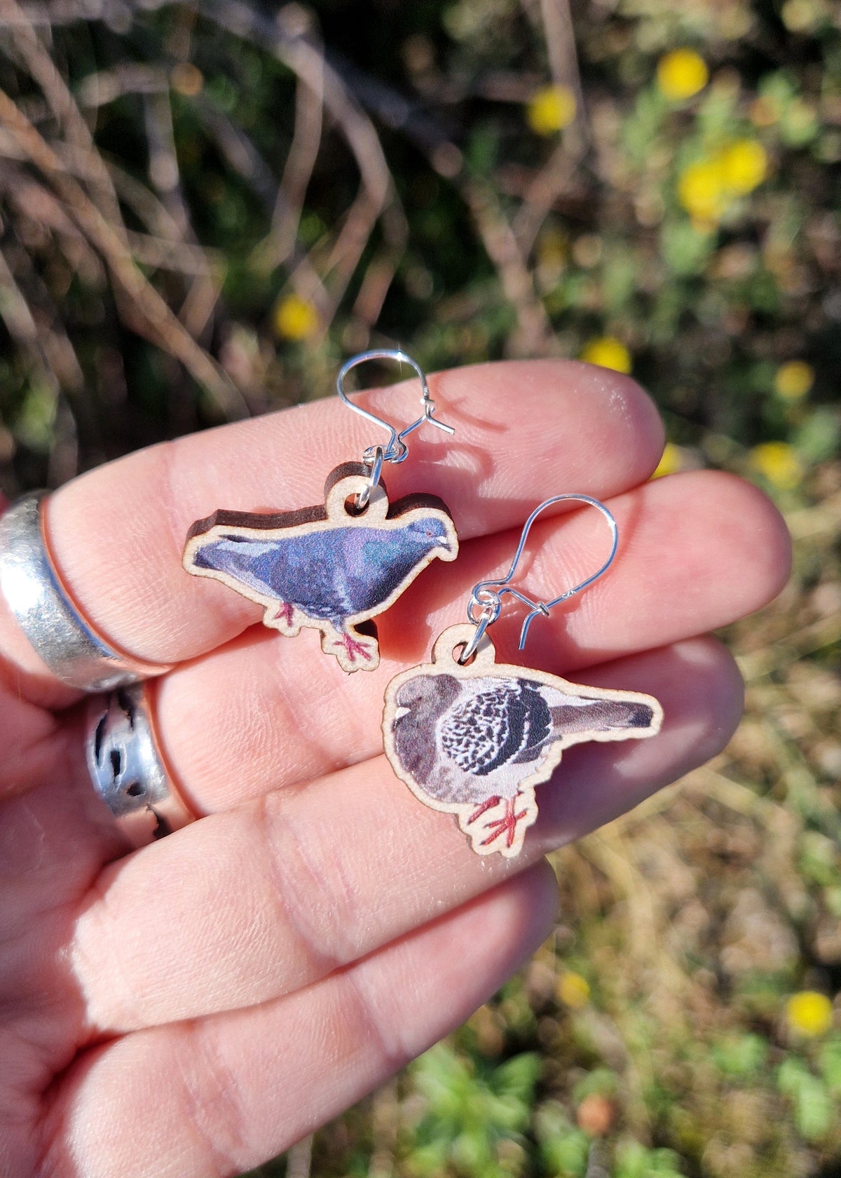 A pair of pigeon earrings with silver findings sit on a white hand wearing silver jewellery above an out of focus yellow flowering plant.