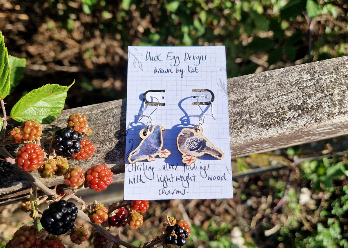 A pair of pigeon earrings sit on a white backing card with the words 'Duck Egg Designs drawn by Kat' in black handwriting. The backing card sits on a weathered wooden fence next to some blackberries on a bramble.