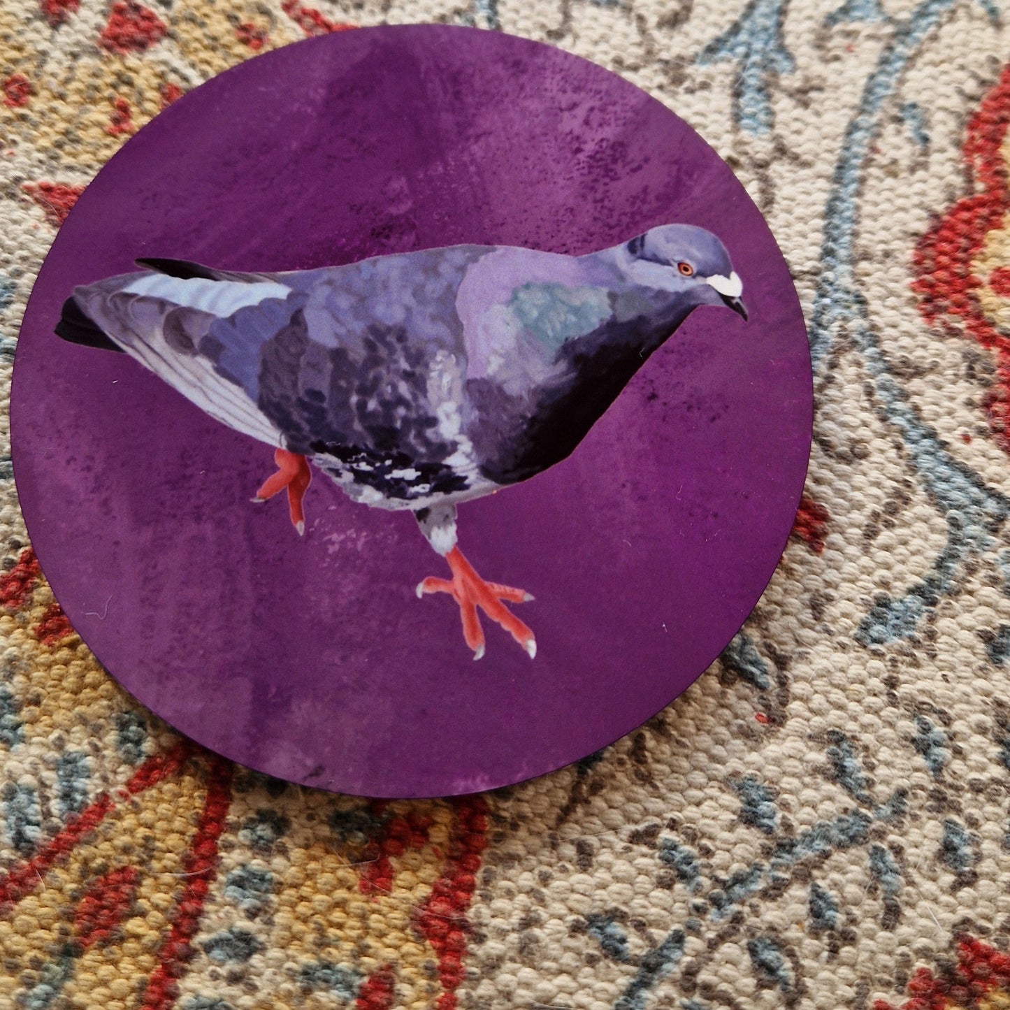 A round purple coaster with a grey pigeon on it sits on a patterned fabric background.