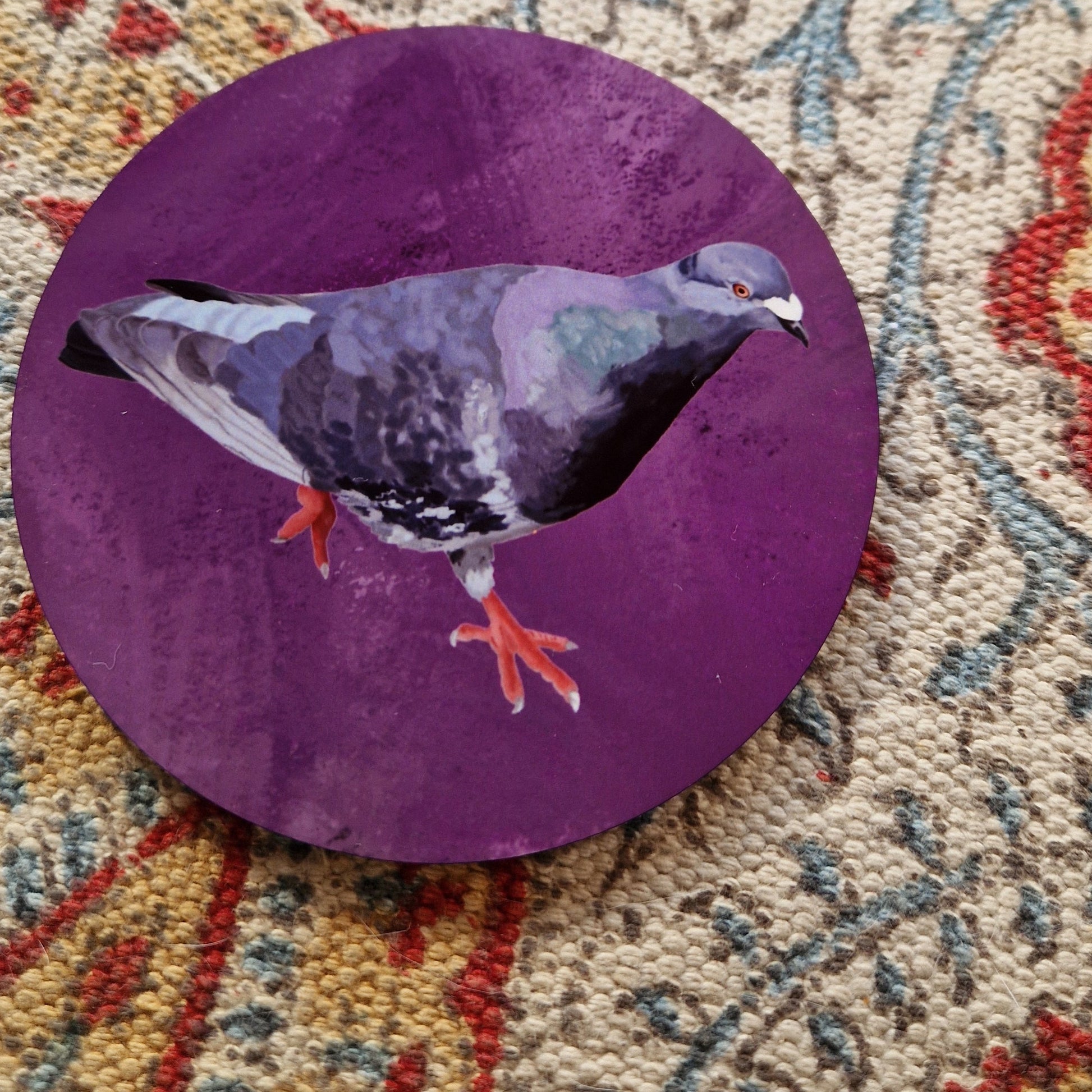 A round purple coaster with a grey pigeon on it sits on a patterned fabric background.
