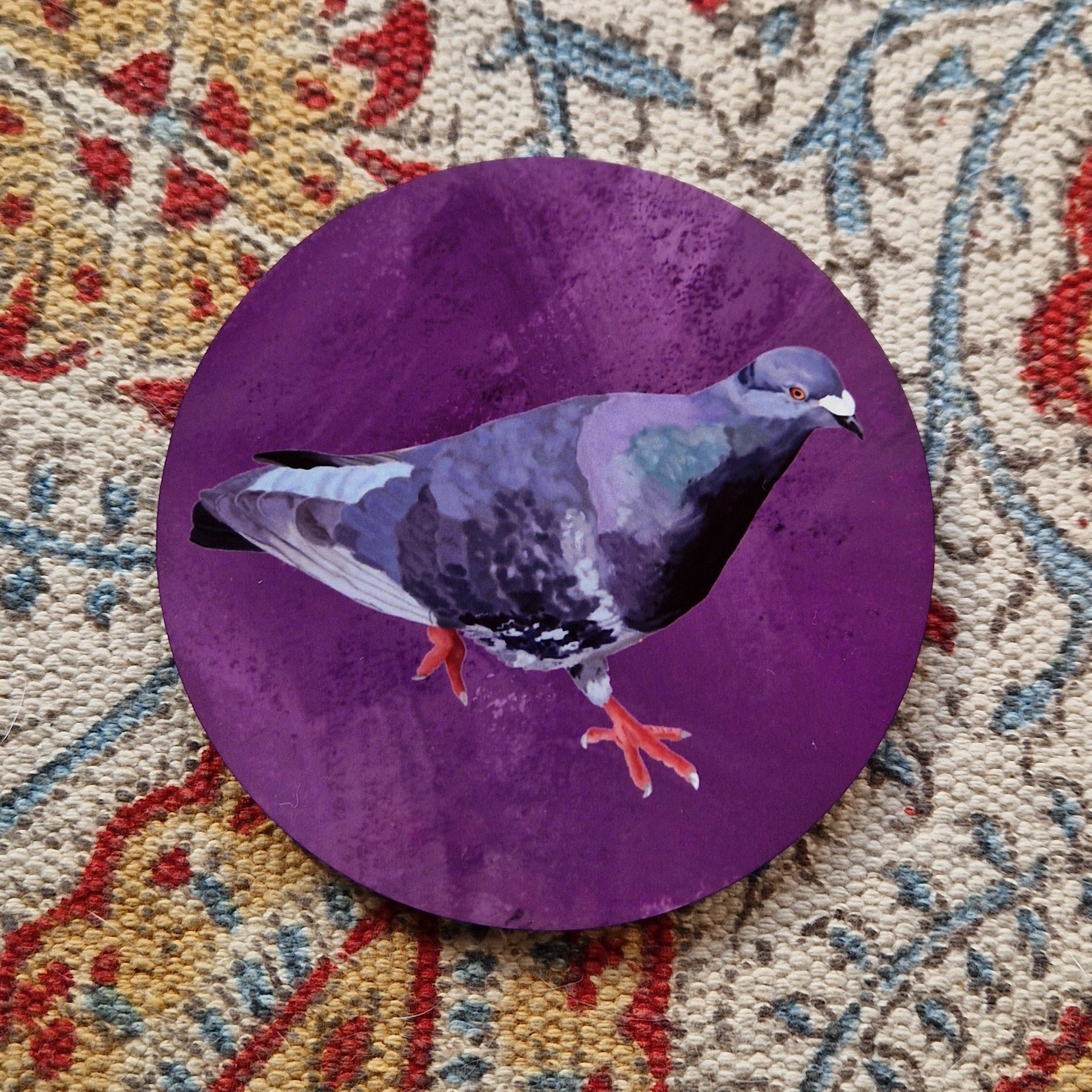A purple coaster featuring a grey, black and blue pigeon with orange feet sits on a patterned fabric background.