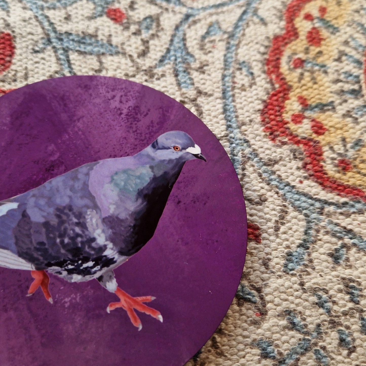A close up of the purple round pigeon coaster showing the detail in the pigeon illustration. Behind the coaster you can see a patterned fabric background.