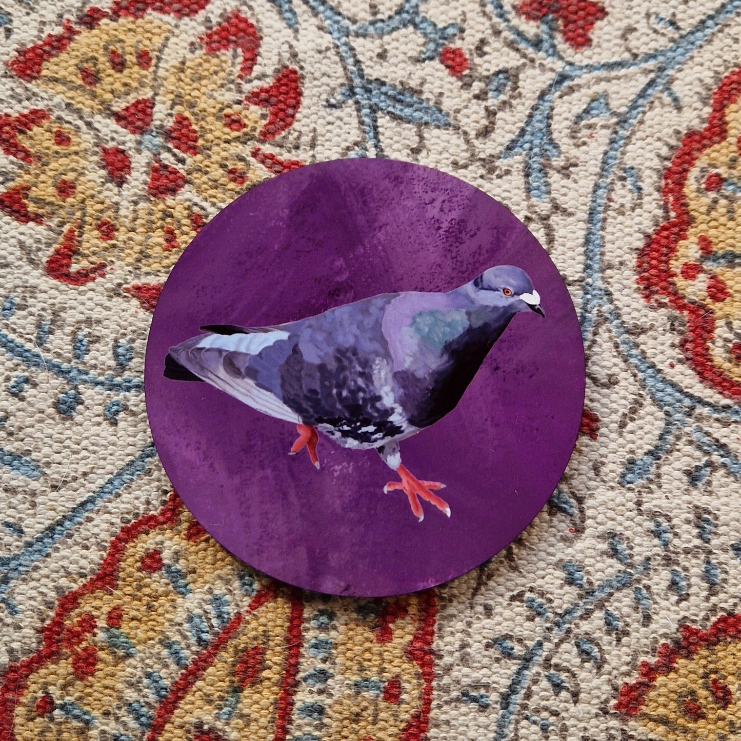A purple coaster featuring a grey, black and blue pigeon with orange feet sits on a patterned fabric background.