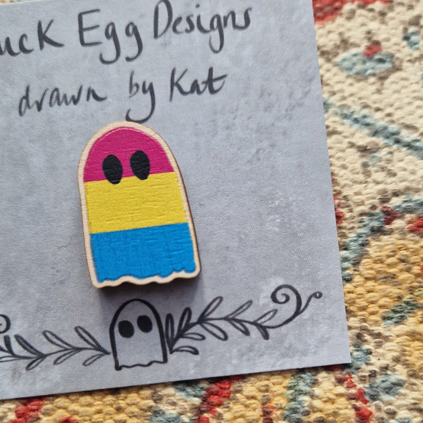 A close up of the pansexual flag ghost pin showing the texture of the wooden pin. The ghost has stripes of pink, yellow and blue; and sits on a grey backing card.