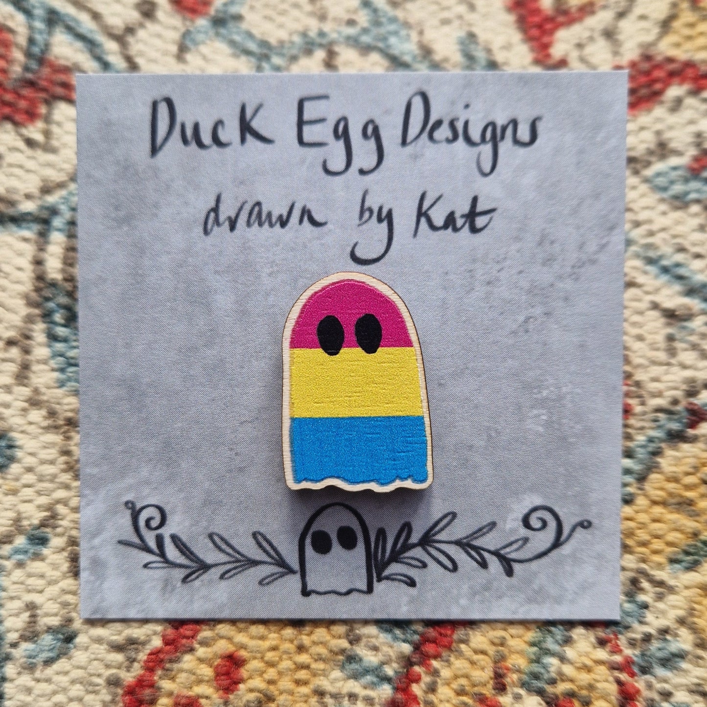 A ghost pin badge in the colours of the pansexual flag (pink, yellow and blue stripes) sits on a grey square backing card  that features the words 'Duck Egg Designs drawn by Kat' in black handwriting. Behind the pin and backing card is a patterned fabric background.