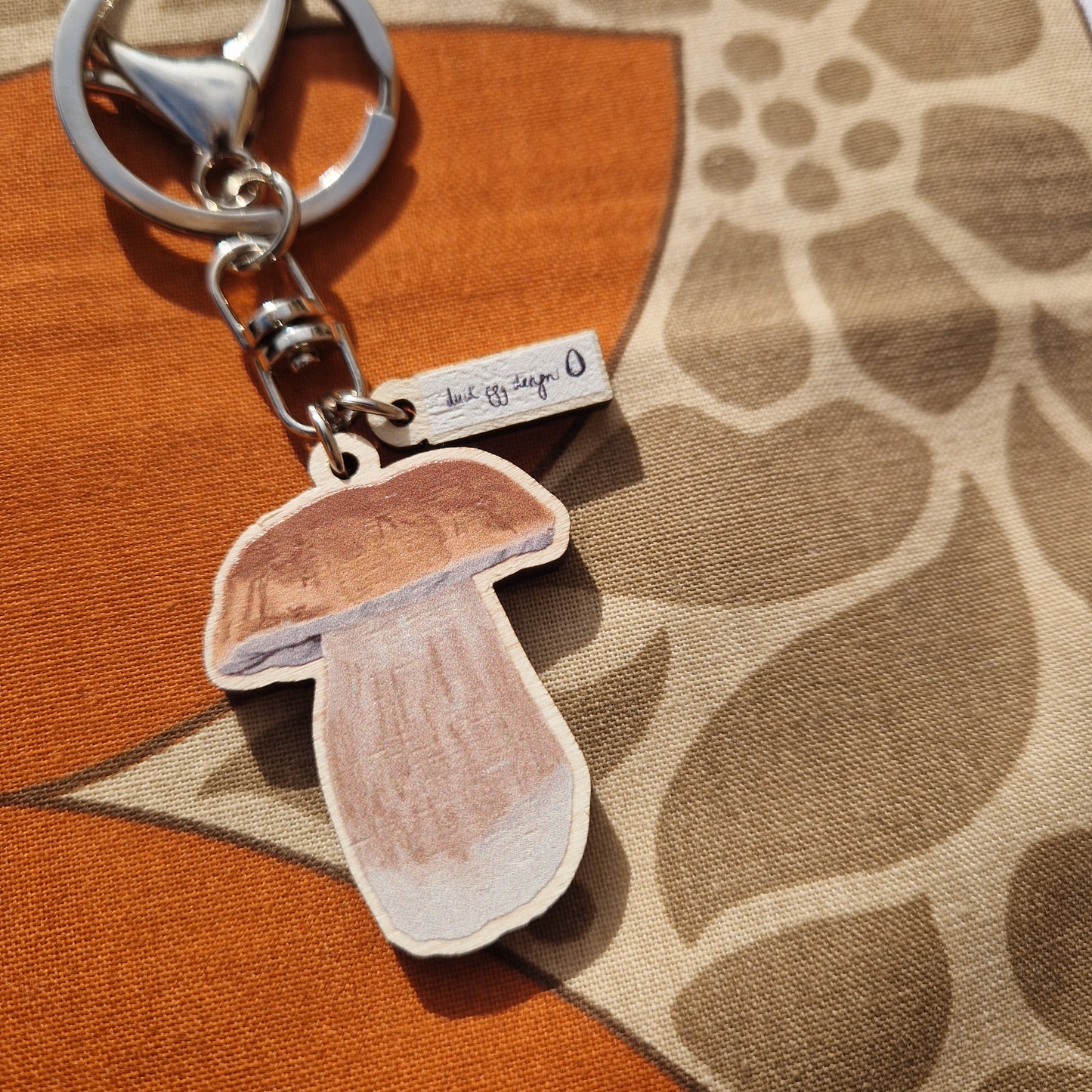 A close up view of the penny bun wooden charm as well as the silver coloured hardware and small white and black Duck Egg Designs logo charm. Behind the keyring is a brown and warm orange fabric.
